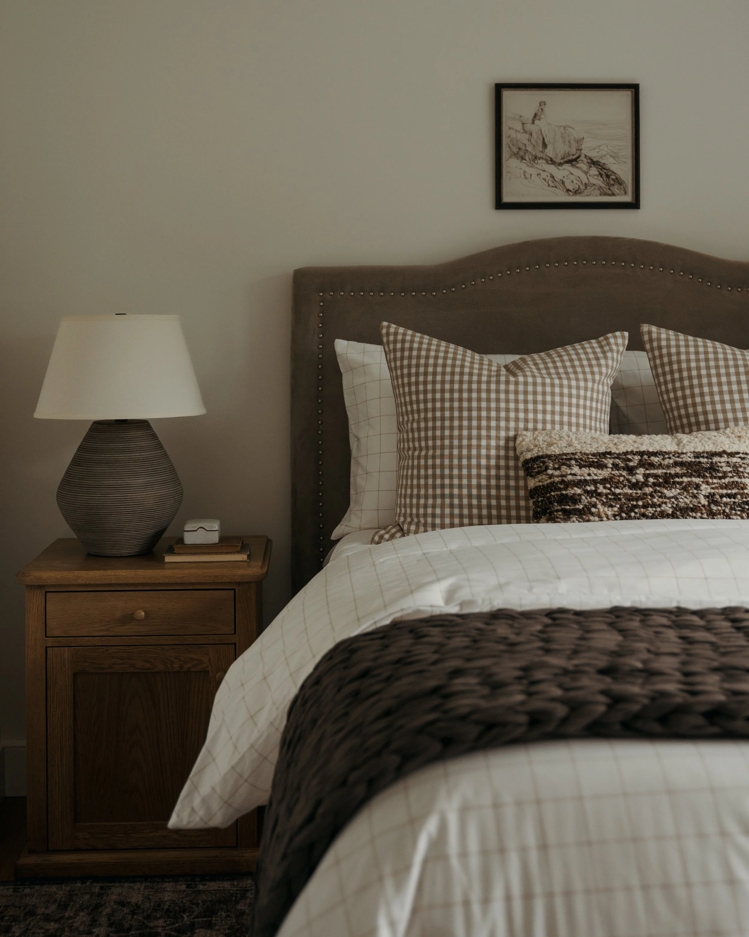 Candace Plotz Design's Project Renewed Roots presents a cozy guest bedroom with a charcoal upholstered headboard featuring nailhead trim. Layered bedding includes white quilted coverlets, brown gingham check pillows, and chunky woven throws in cream 
