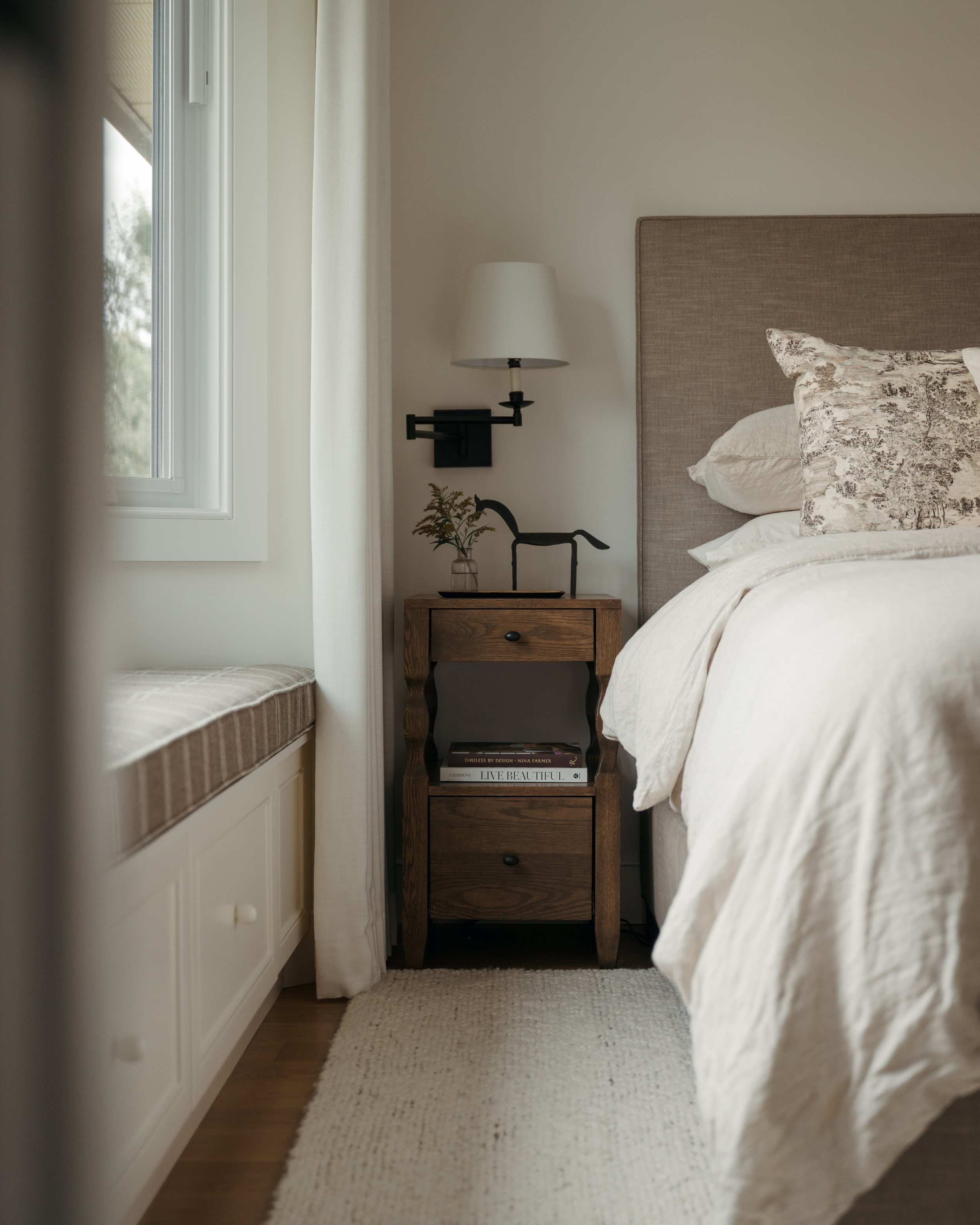 Candace Plotz Design's Project Renewed Roots presents a serene primary bedroom corner with a tall window framing natural light. A walnut nightstand with two drawers holds a black swing-arm sconce, small bronze sculpture, and delicate dried botanicals