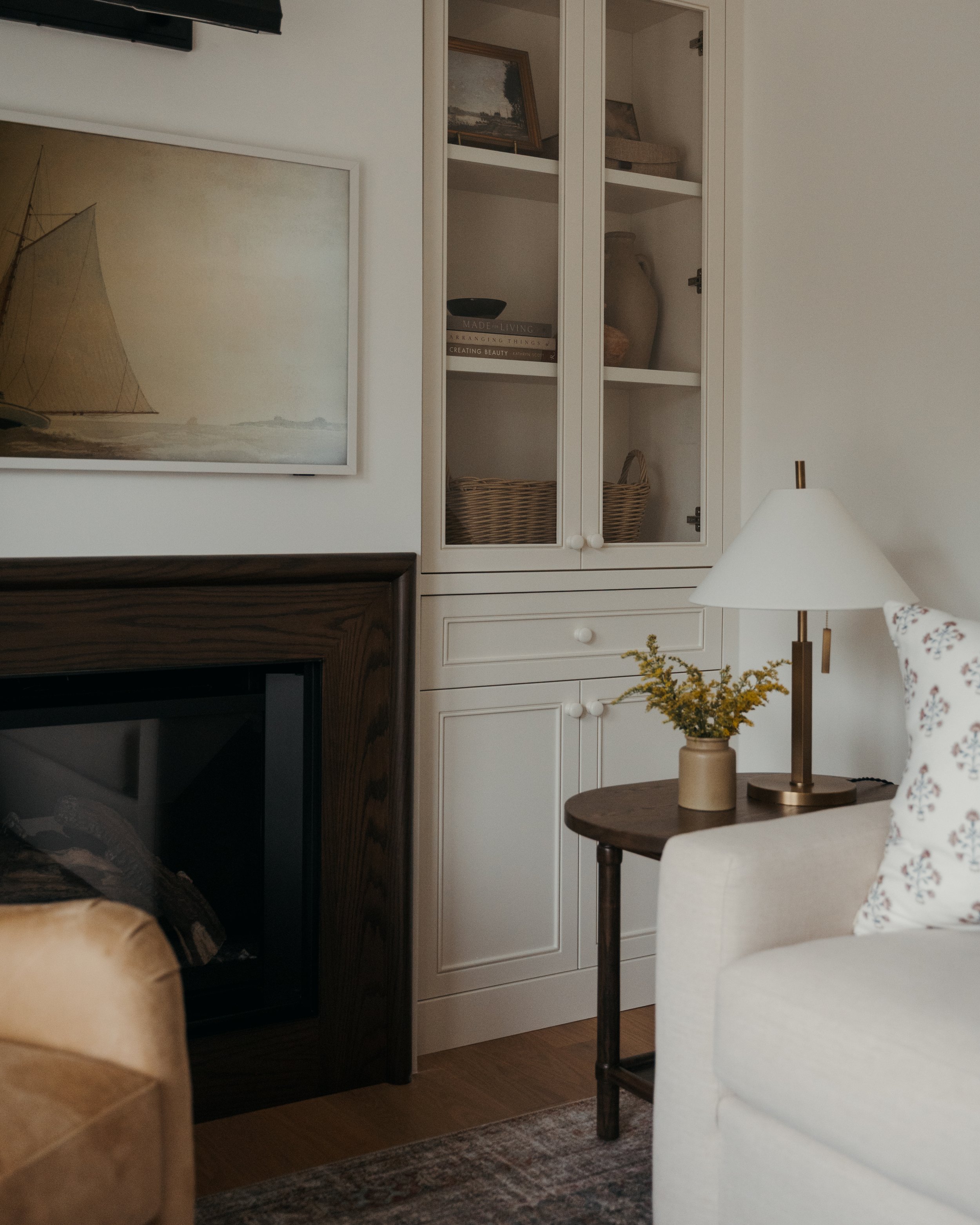 A living room built-in in Candace Plotz Design's Project Renewed Roots flanks a dark wood-framed fireplace with black insert. White glass-front cabinets display curated books, woven baskets, and pottery, while lower drawers feature white knobs. A vin