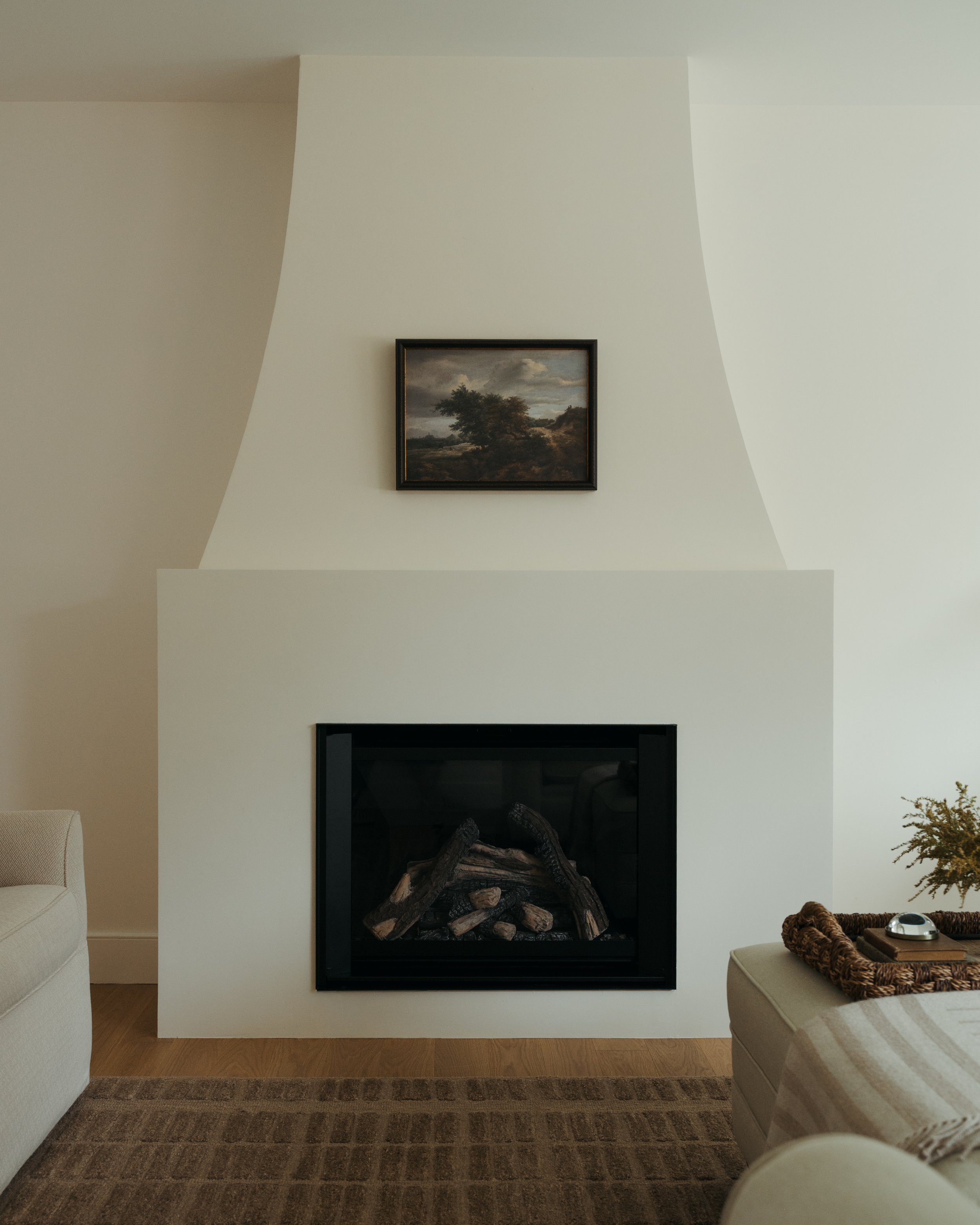An intimate dining room corner in Candace Plotz Design's Project Renewed Roots highlights textural details including linen slipcovered chairs with pleated skirts, honey-toned oak flooring, and crisp white wainscoting. The moody landscape painting wit
