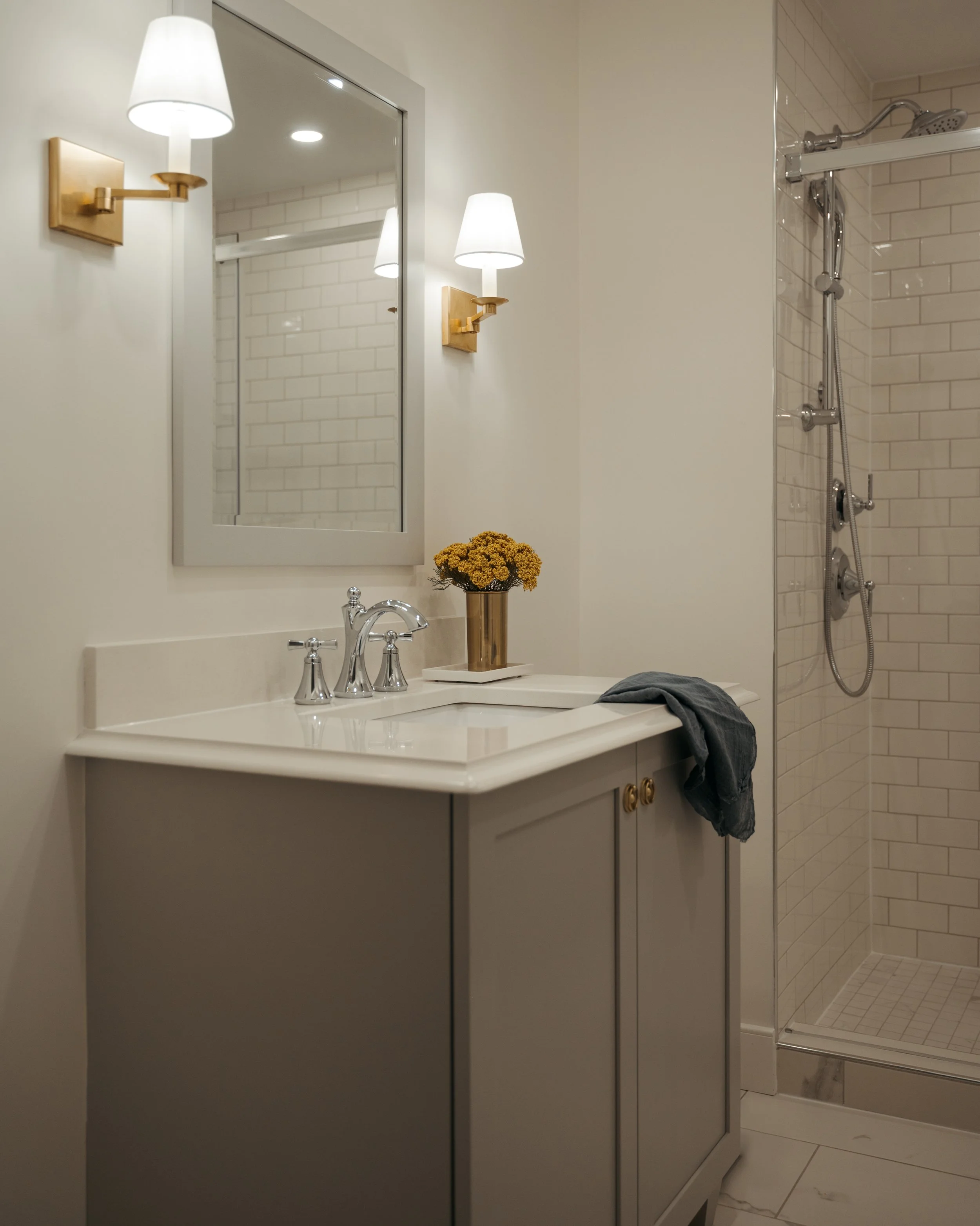 Candace Plotz Design's Project Renewed Roots features a compact bathroom with a taupe-gray vanity cabinet topped with white quartz and polished chrome traditional faucet. Brass sconces with white shades flank a white-framed mirror reflecting classic 