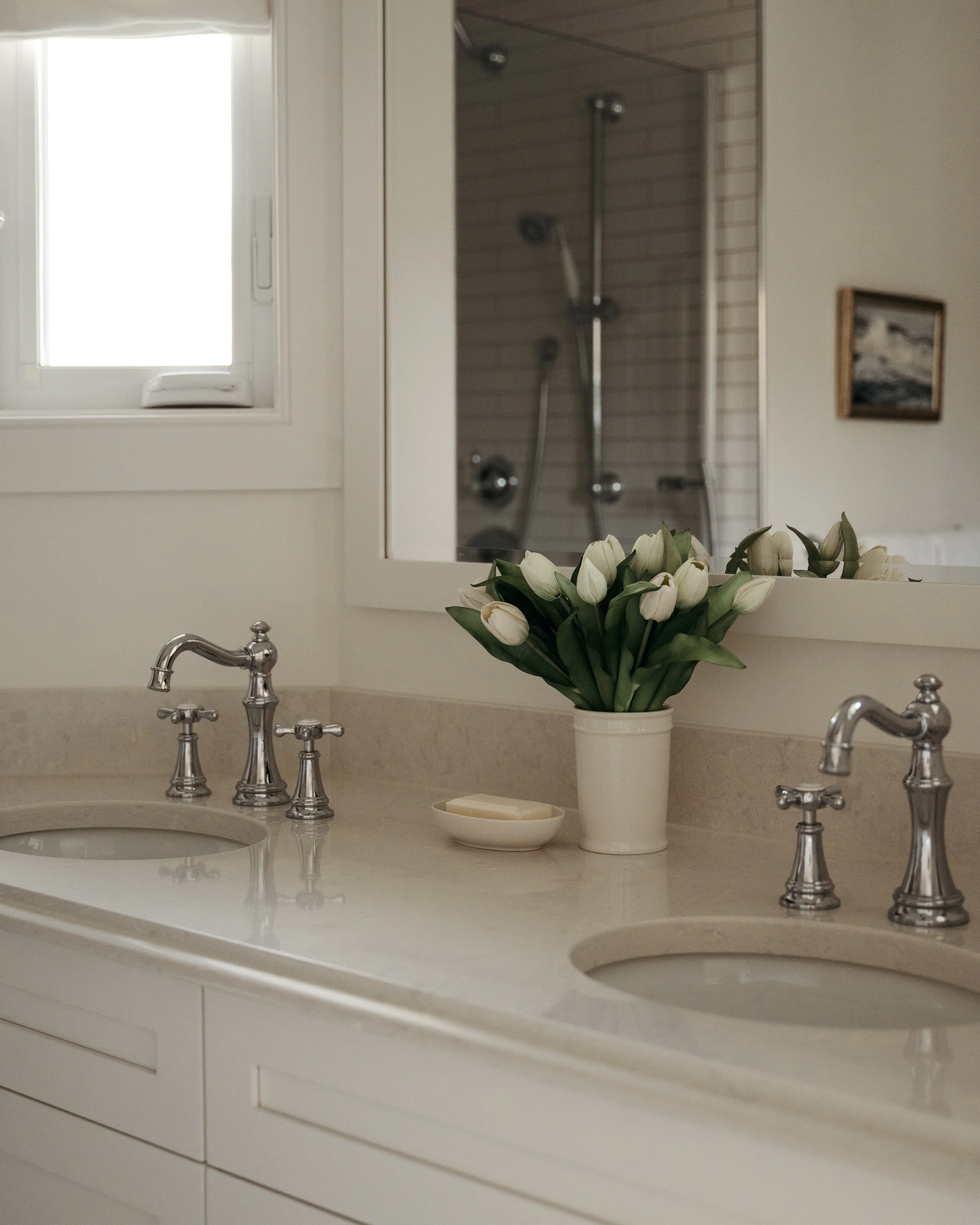 A classic bathroom vanity in Candace Plotz Design's Project Renewed Roots features dual undermount sinks with polished chrome traditional faucets and a honed marble countertop. White tulips in a ceramic vase provide fresh elegance between the sinks, 