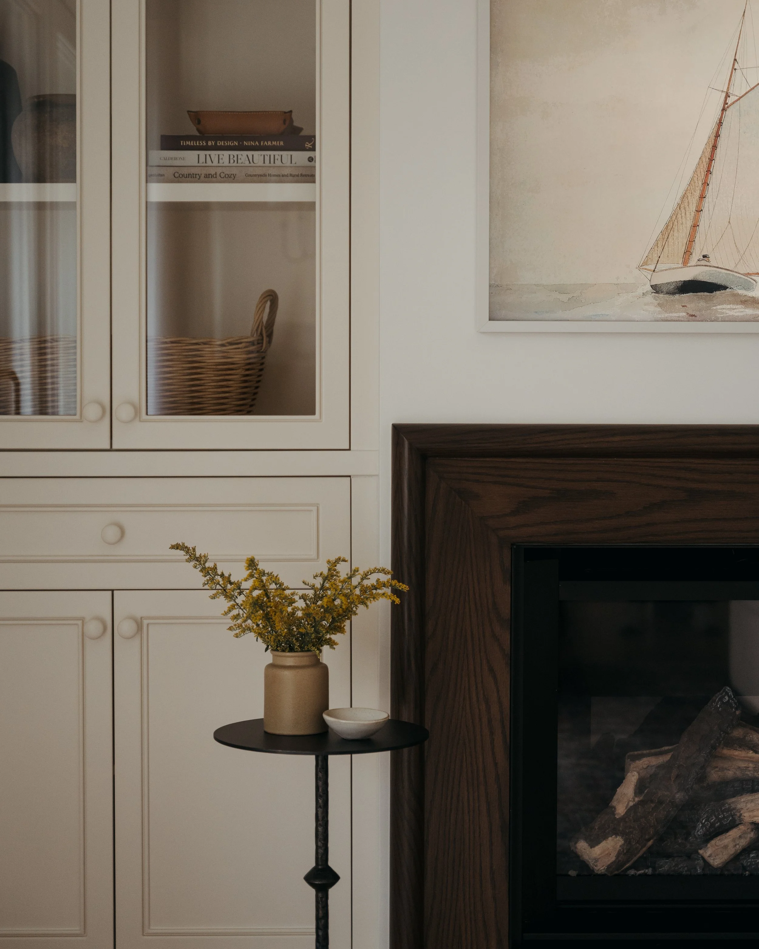 Candace Plotz Design's Project Renewed Roots showcases fireplace built-in storage detail with white glass-front cabinets revealing stacked design books and a woven basket. A vintage sailboat painting in cream tones hangs adjacent to the dark wood fir