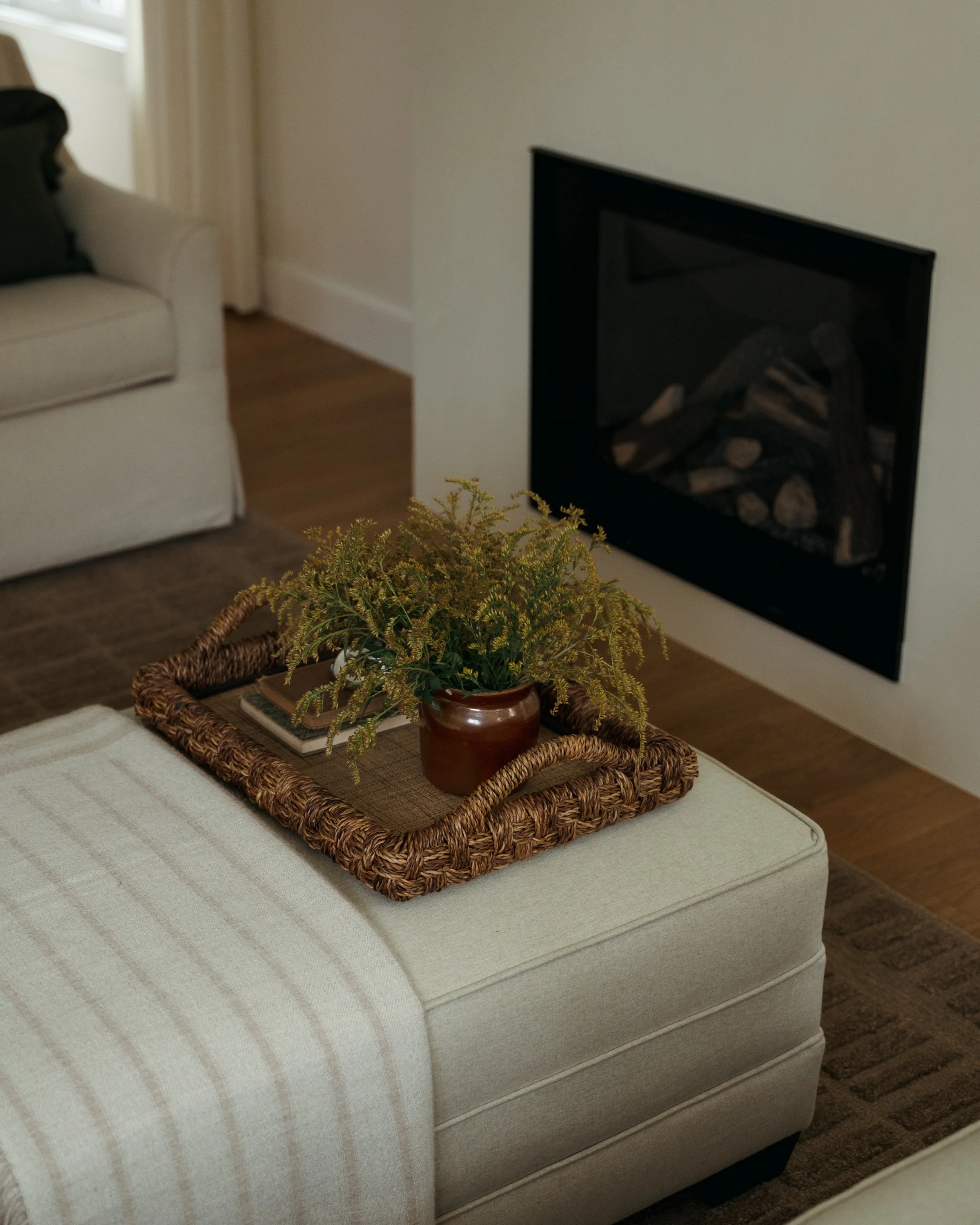 A cozy living room detail in Candace Plotz Design's Project Renewed Roots highlights a woven seagrass tray styled atop a striped cream ottoman. The tray holds a terracotta pot with cascading greenery and stacked books, positioned before the minimalis