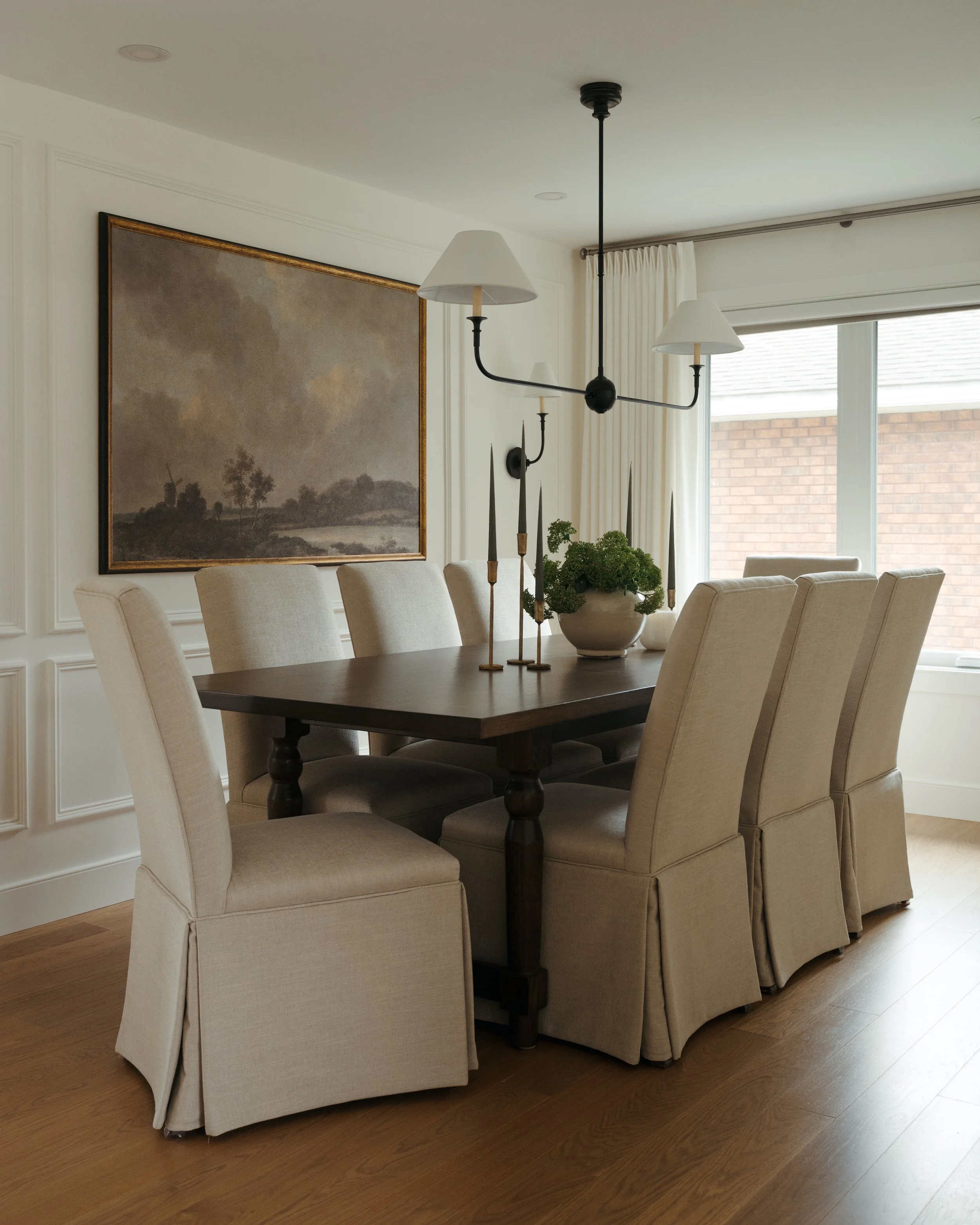 A traditional dining room in Candace Plotz Design's Project Renewed Roots features a dark wood pedestal table with linen-skirted parsons chairs arranged for eight. An oversized landscape painting with gold frame provides focal drama behind a modern t