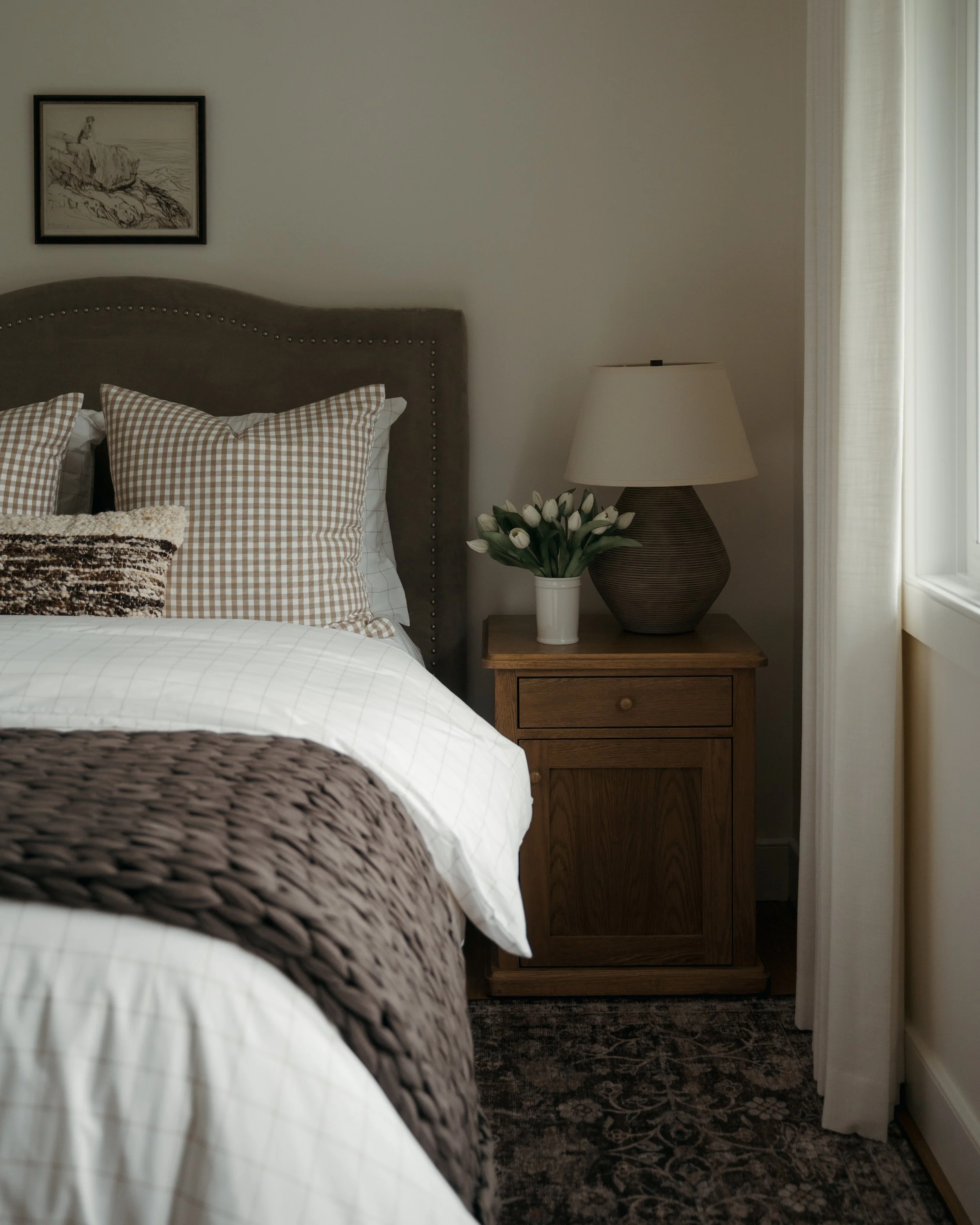 In Candace Plotz Design's Project Renewed Roots, a peaceful bedroom vignette captures golden afternoon light. The charcoal linen headboard with brass nailheads anchors layered bedding in gingham checks and neutral tones, while a walnut nightstand dis