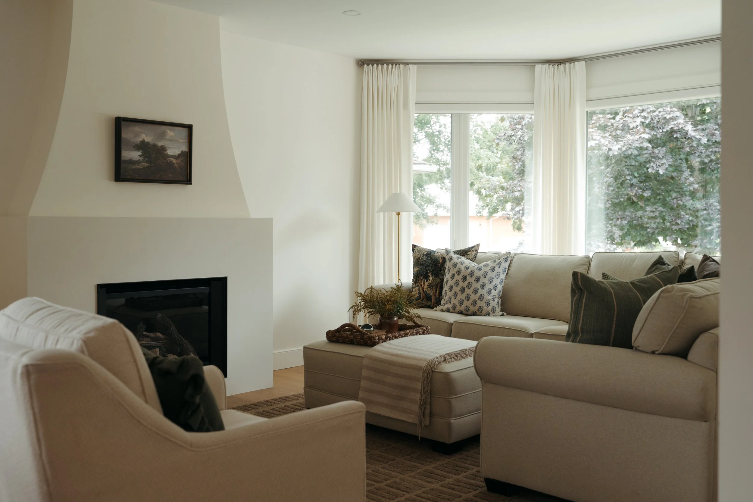  In Candace Plotz Design's Project Renewed Roots, a serene living room showcases a sculptural white plaster fireplace surround with black firebox insert. Taupe linen sofas flank a woven ottoman coffee table topped with dried botanicals. A classical l