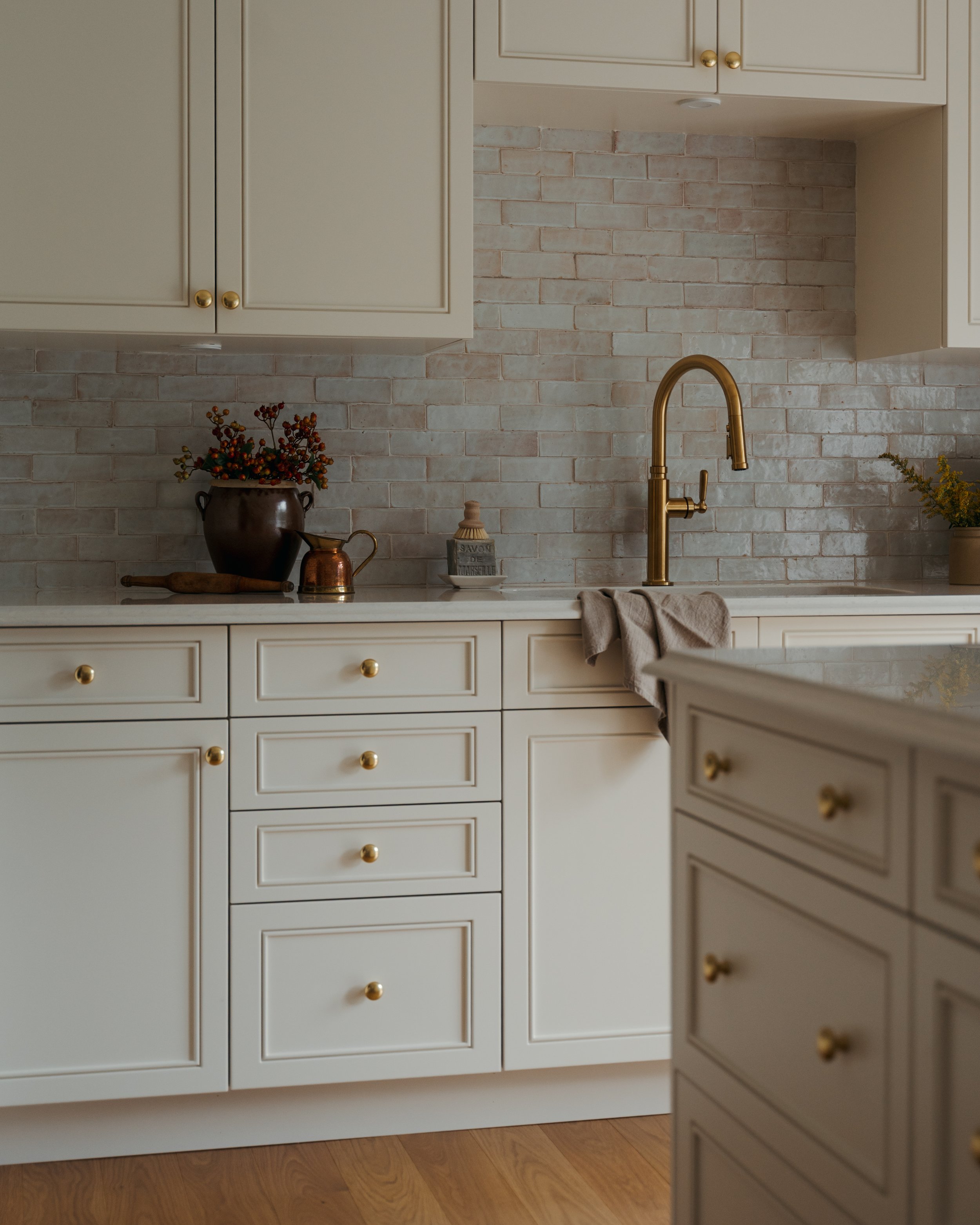 An intimate kitchen corner in Candace Plotz Design's Project Renewed Roots reveals the layered textures of ivory cabinetry with brass knobs against handmade terracotta brick backsplash. The white marble countertop showcases artisanal styling with a d