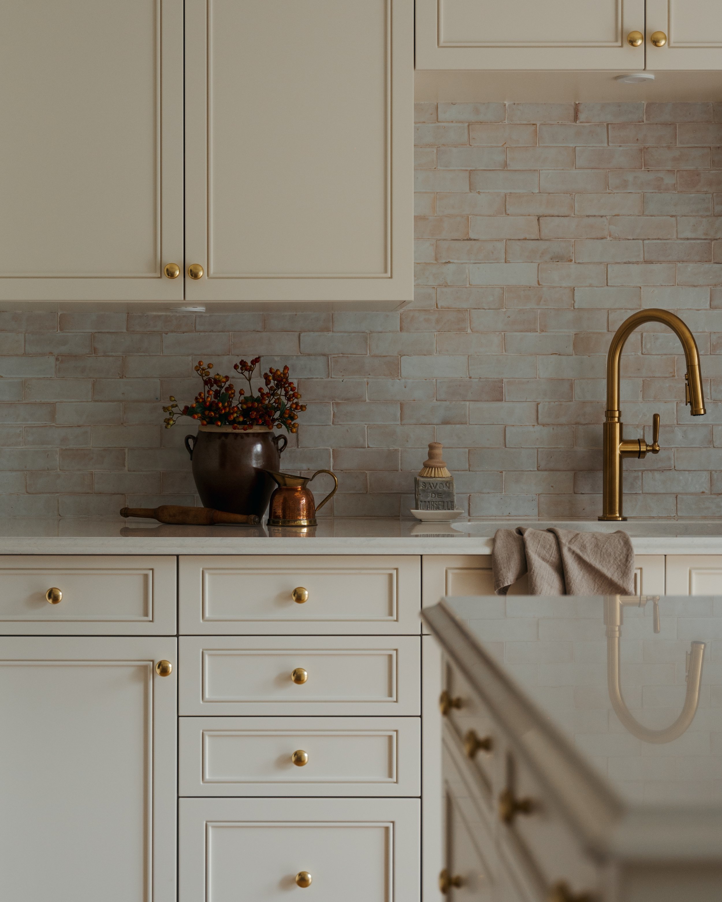 Candace Plotz Design's Project Renewed Roots presents a refined kitchen workspace with floor-to-ceiling ivory cabinetry featuring brass hardware and handmade terracotta brick backsplash. White marble countertops and a high-arc brass faucet anchor the