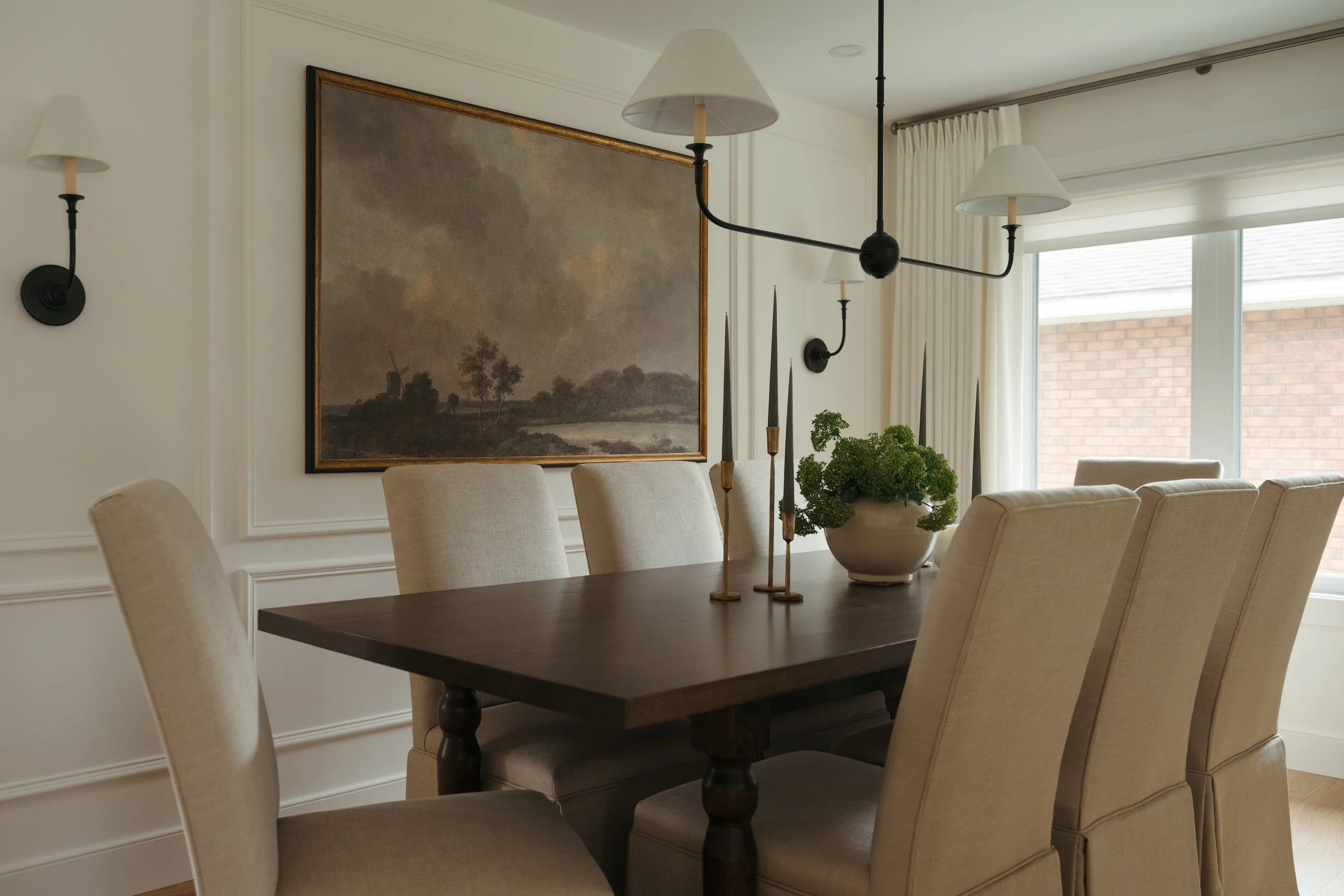 Candace Plotz Design's Project Renewed Roots features an elegant dining room with a dark wood trestle table surrounded by eight linen-slipcovered chairs beneath a black three-arm chandelier with white shades. A large landscape painting in a gilt fram