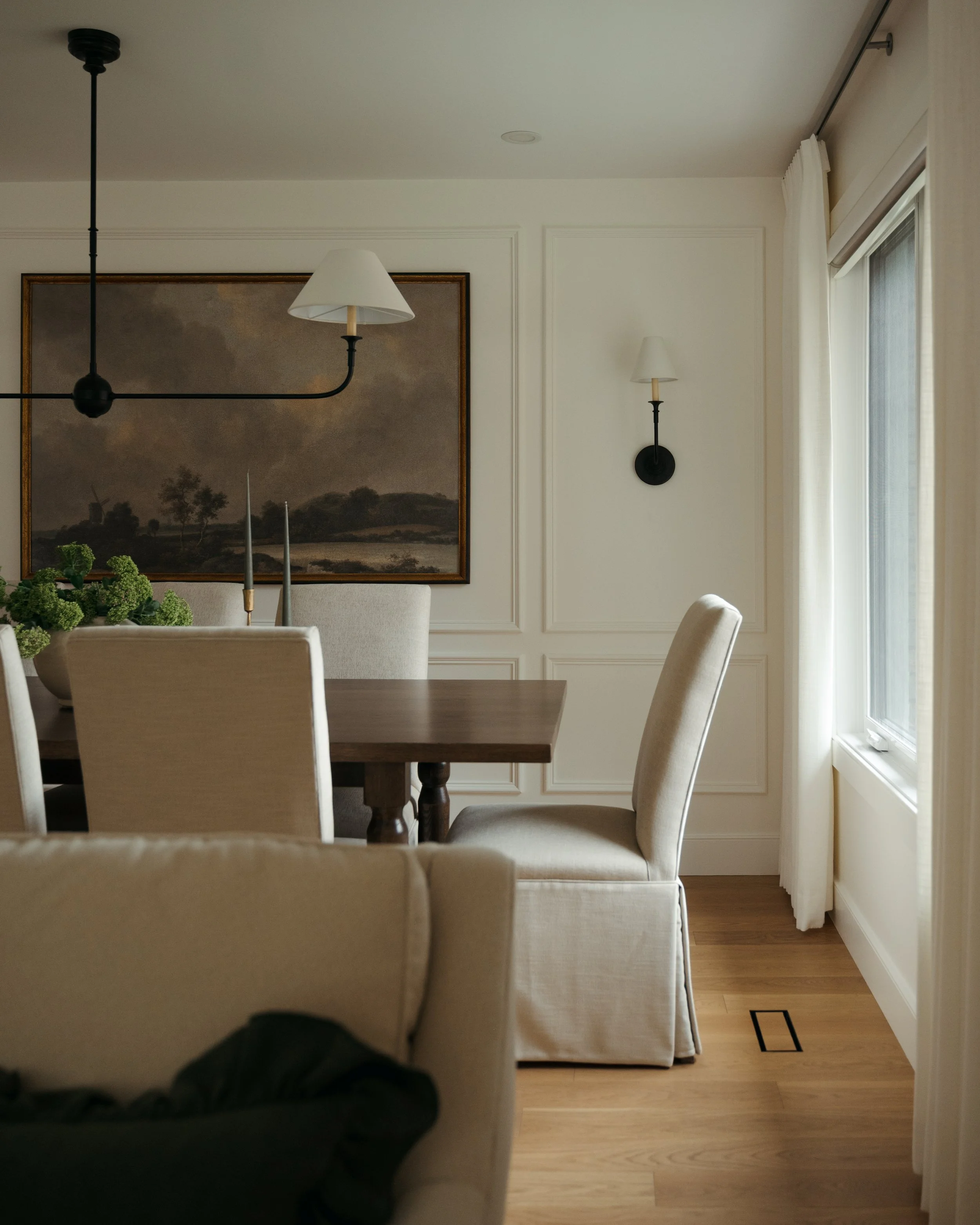 An intimate dining room corner in Candace Plotz Design's Project Renewed Roots highlights textural details including linen slipcovered chairs with pleated skirts, honey-toned oak flooring, and crisp white wainscoting. The moody landscape painting wit