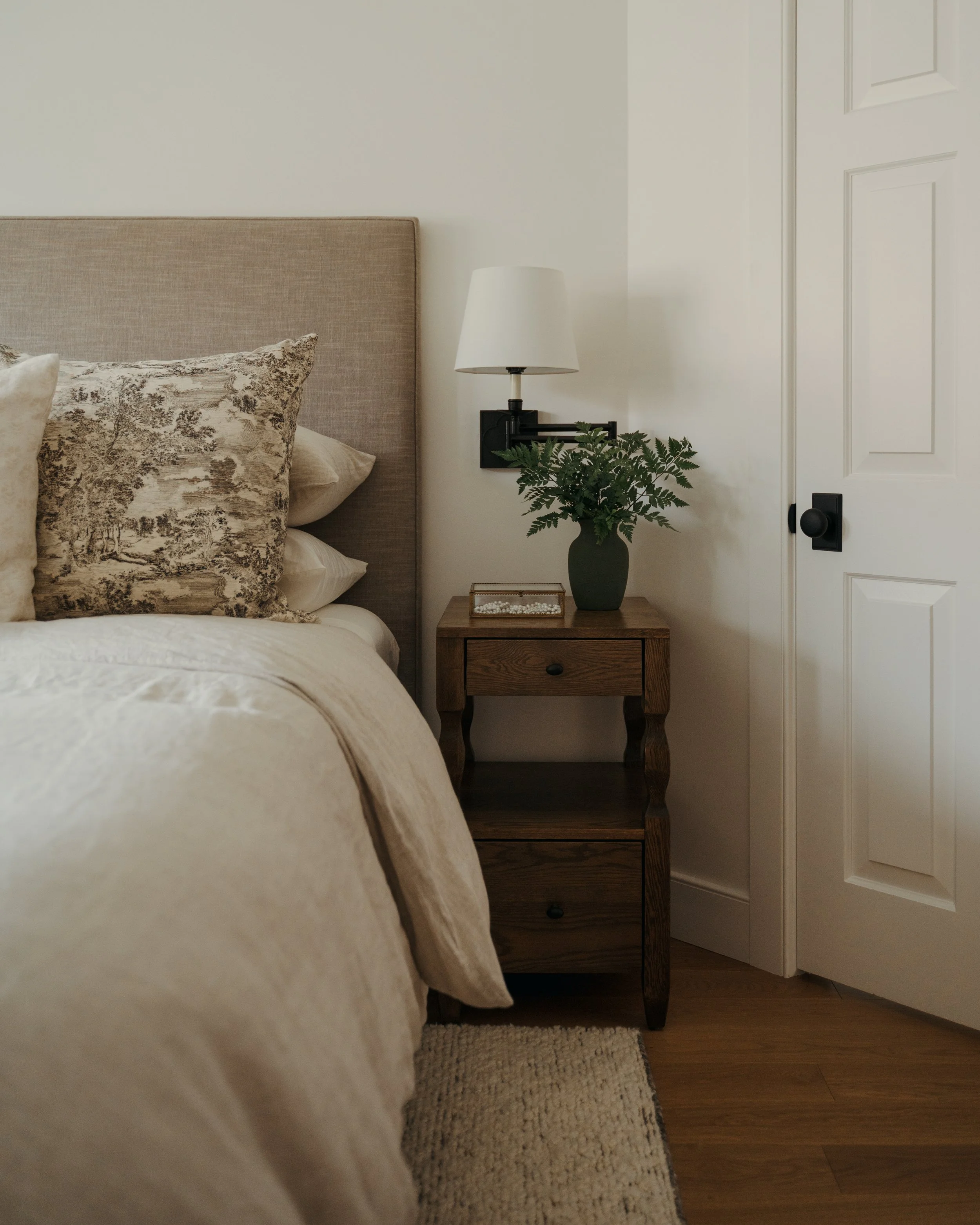  In Candace Plotz Design's Project Renewed Roots, a tranquil bedroom detail reveals a walnut two-drawer nightstand between the taupe linen headboard and white five-panel door with matte black hardware. A black swing-arm wall sconce with white shade i