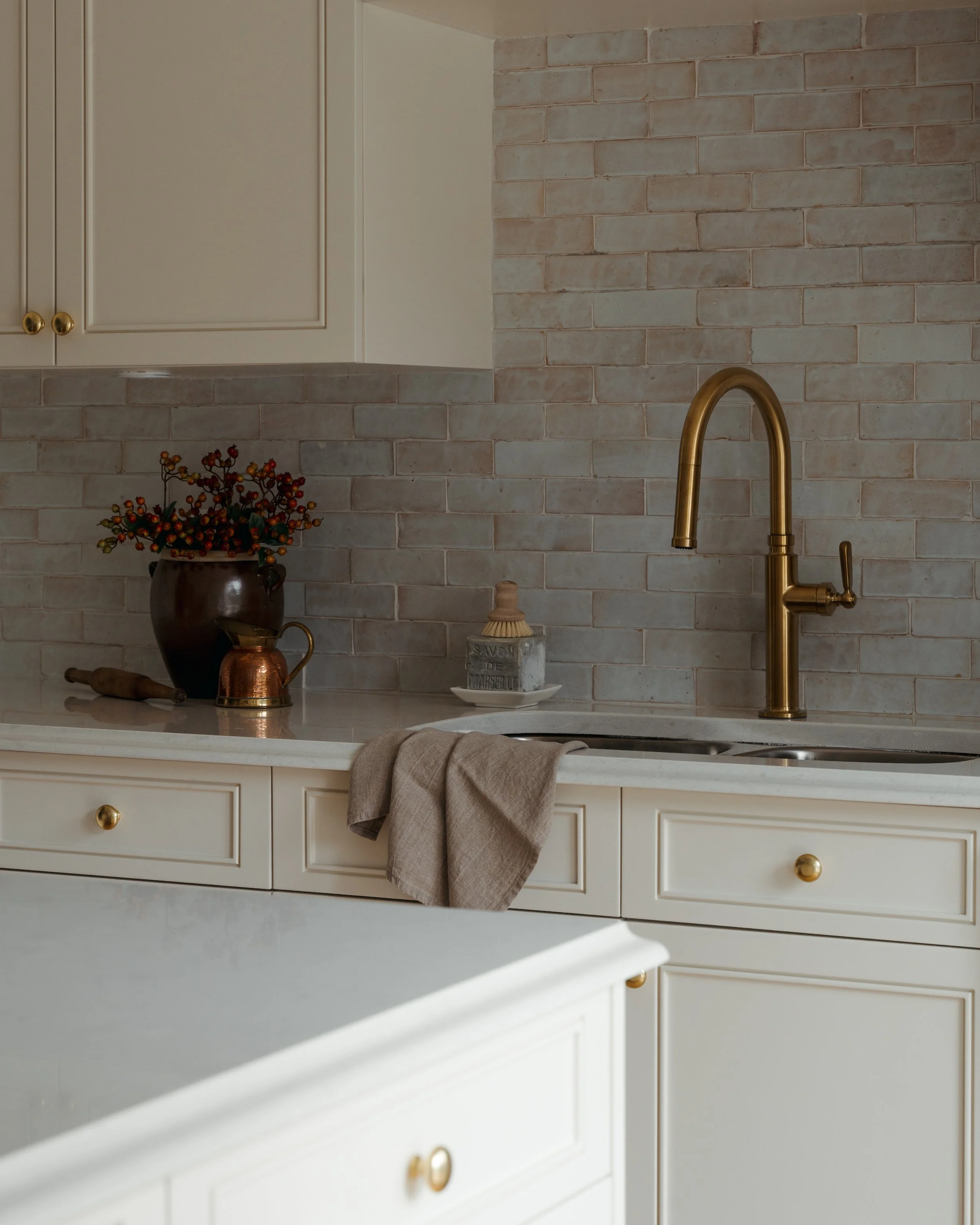 Candace Plotz Design's Project Renewed Roots highlights kitchen sink details with a gooseneck brass faucet against soft terracotta brick backsplash. White marble countertops display curated vintage finds including a dark ceramic jug with autumn botan
