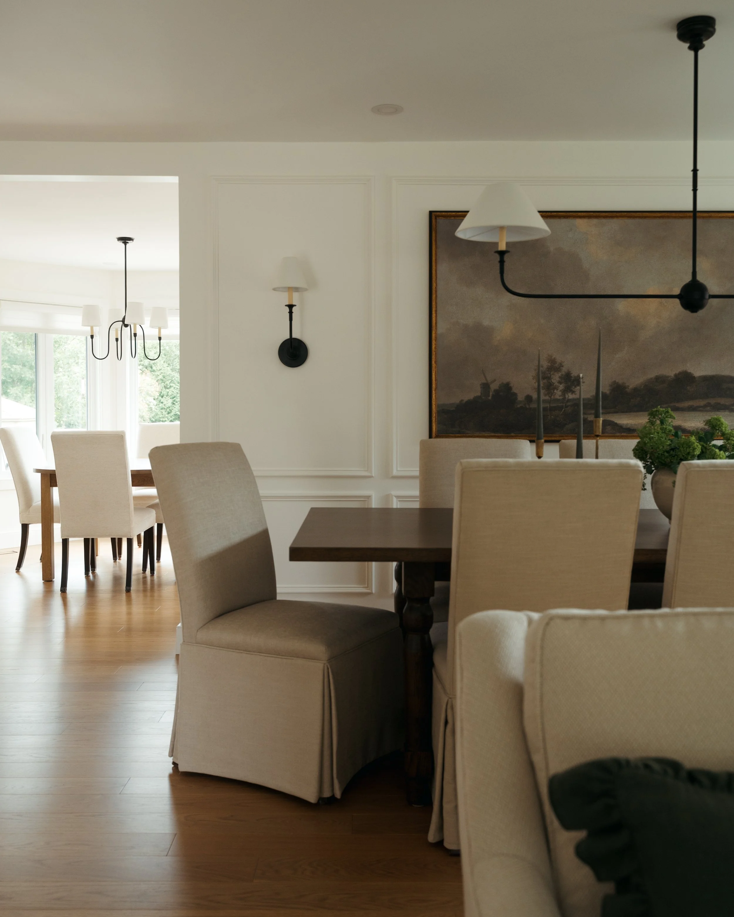 Candace Plotz Design's Project Renewed Roots showcases an open-concept dining area revealing layered living spaces through a classical doorway with panel molding. A dark wood dining table with slipcovered chairs anchors the foreground beneath asymmet