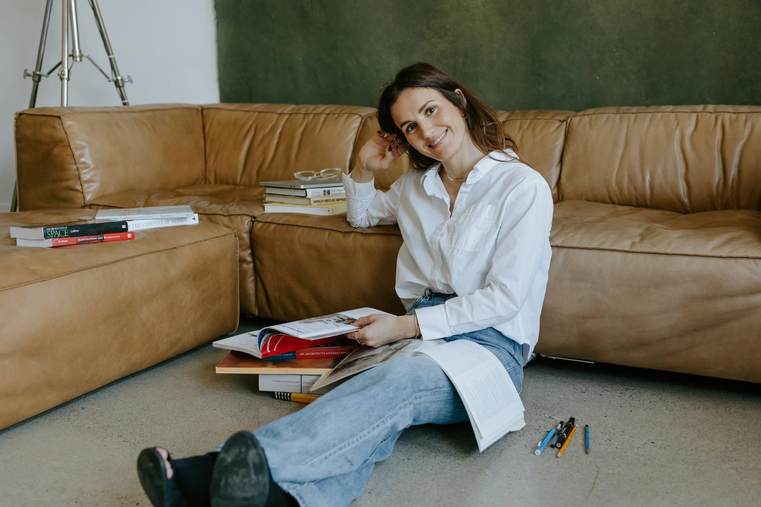 A woman with shoulder-length dark hair, wearing a white shirt and blue jeans, sitting on the floor in front of a tan leather sofa, smiling at the camera. She has an open book in her lap, surrounded by various other books and pens on the floor. There are more books stacked on the sofa behind her, and a green wall with a blackboard or dark panel behind her. There is also a tripod stand visible in the background.
