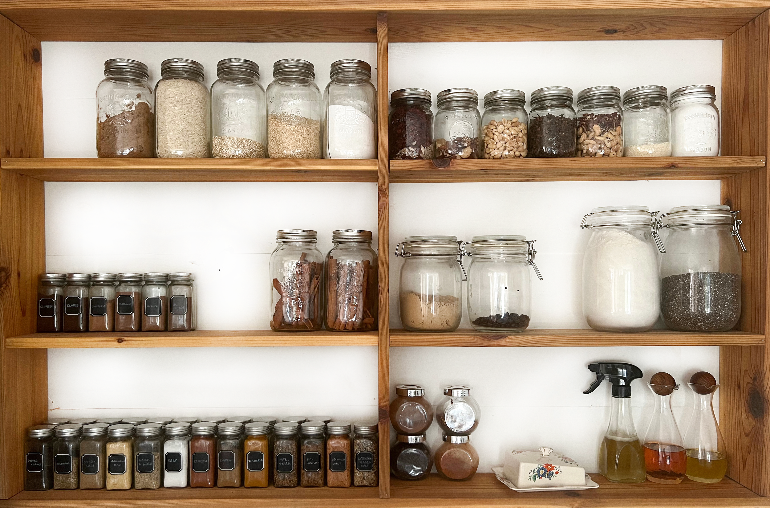 Wooden kitchen shelf filled with glass jars containing spices, herbs, grains, dried fruits, and seasonings. Some jars have chalkboard labels with spice names.
