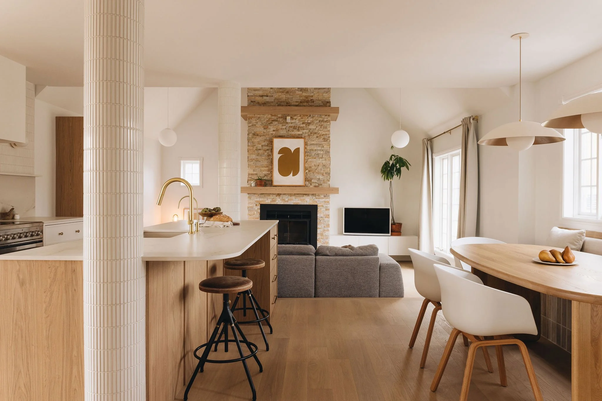 Intérieur d'un salon et d'une cuisine modernes, avec cheminée en pierre, canapé gris, télévision, table en bois, chaises blanches, lumière naturelle, plantes, et décoration minimaliste.