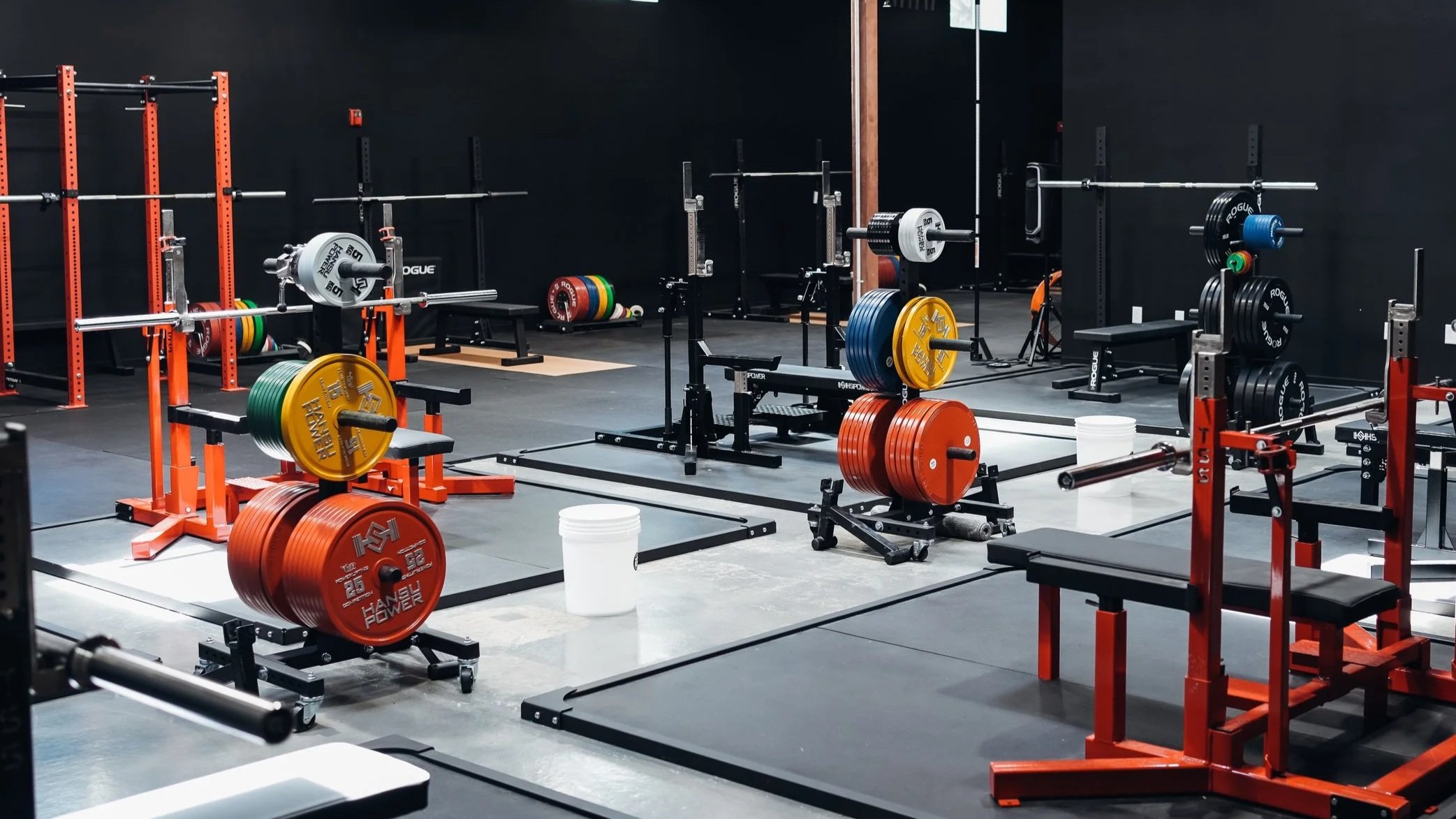 A gym with weightlifting equipment, including barbells, weight plates of various colors, squat racks, benches, and lifting platforms, set against black walls.