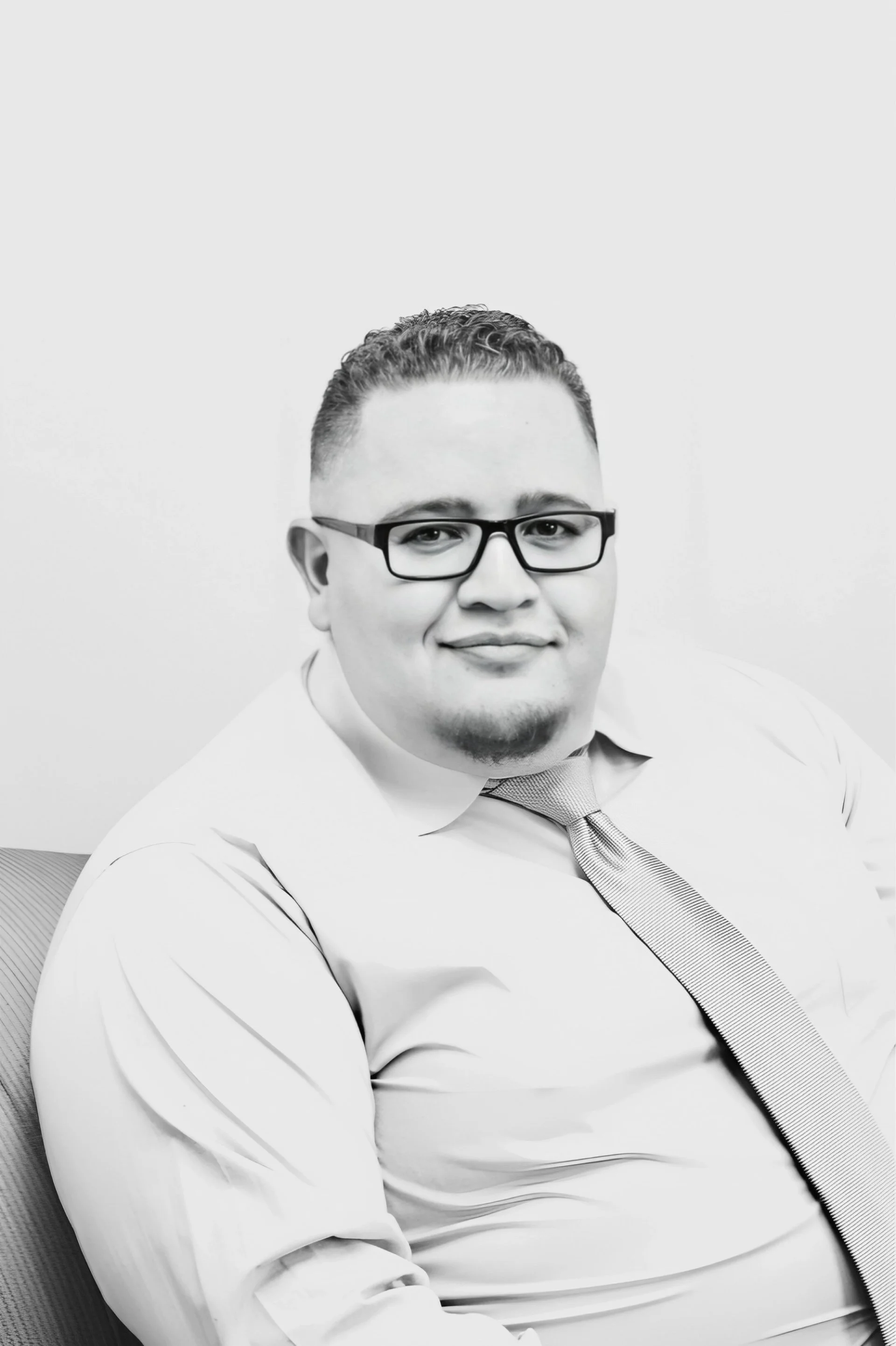 Black and white portrait of a man with glasses, wearing a dress shirt and tie, seated and smiling.
