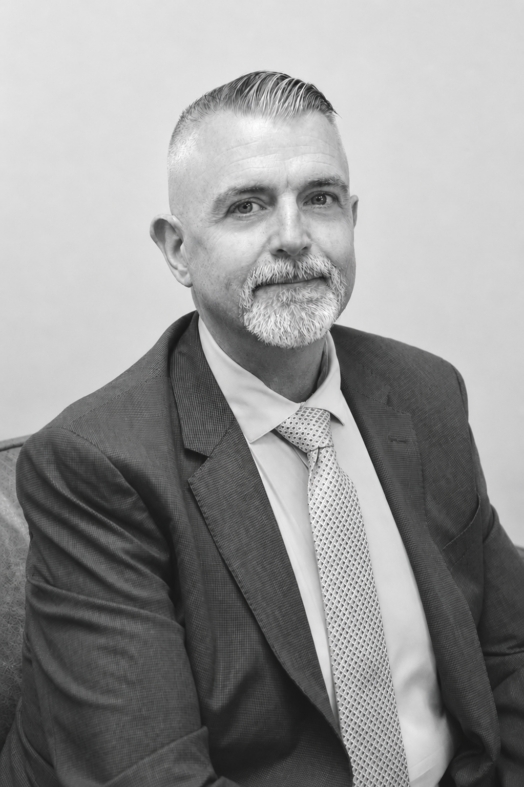 A black-and-white photo of a middle-aged man with a beard and mustache, wearing a suit, tie, and white shirt, sitting on a chair against a plain wall background.