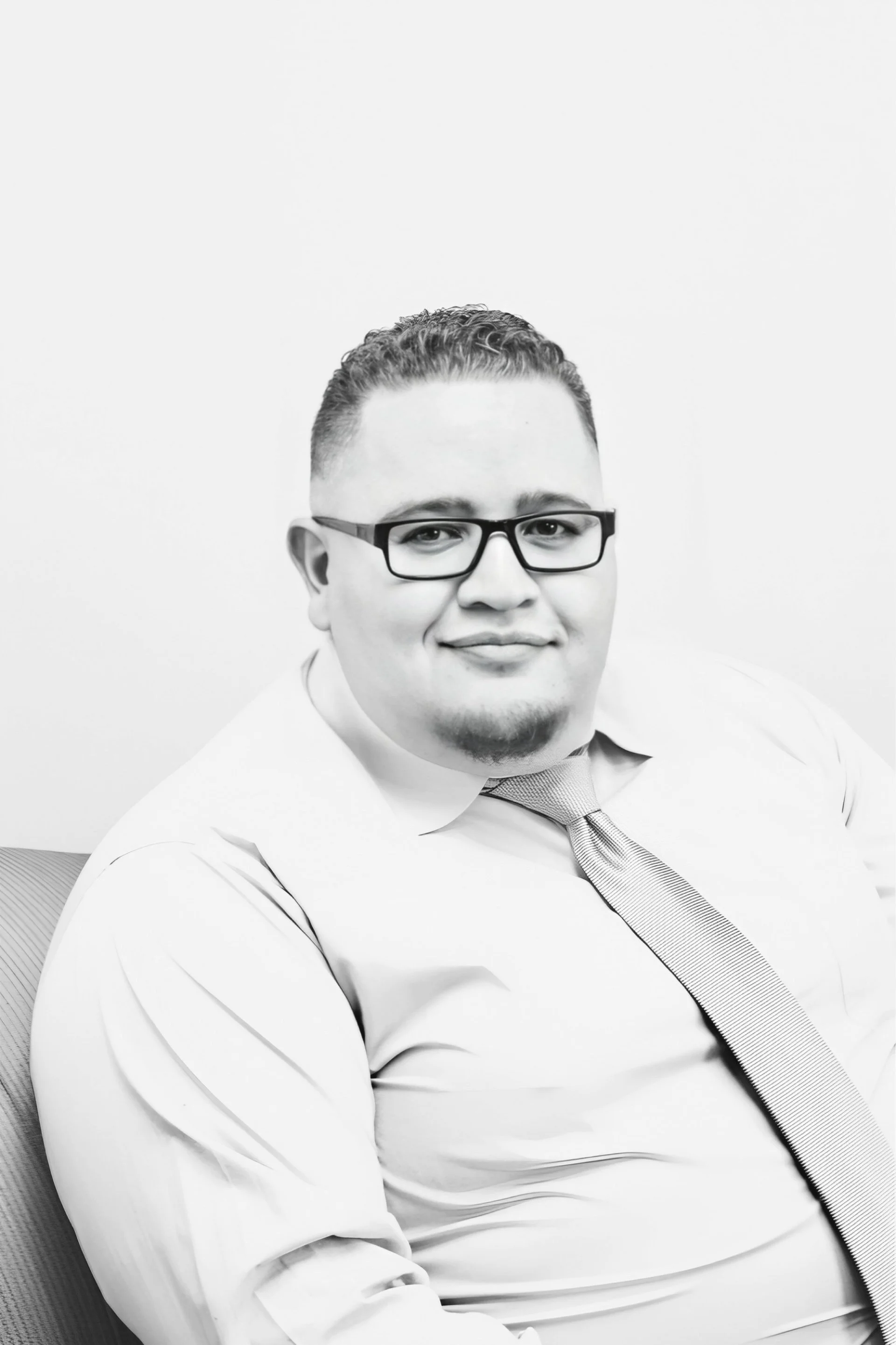 Black and white portrait of a person wearing glasses, a dress shirt, and a tie, sitting on a chair.