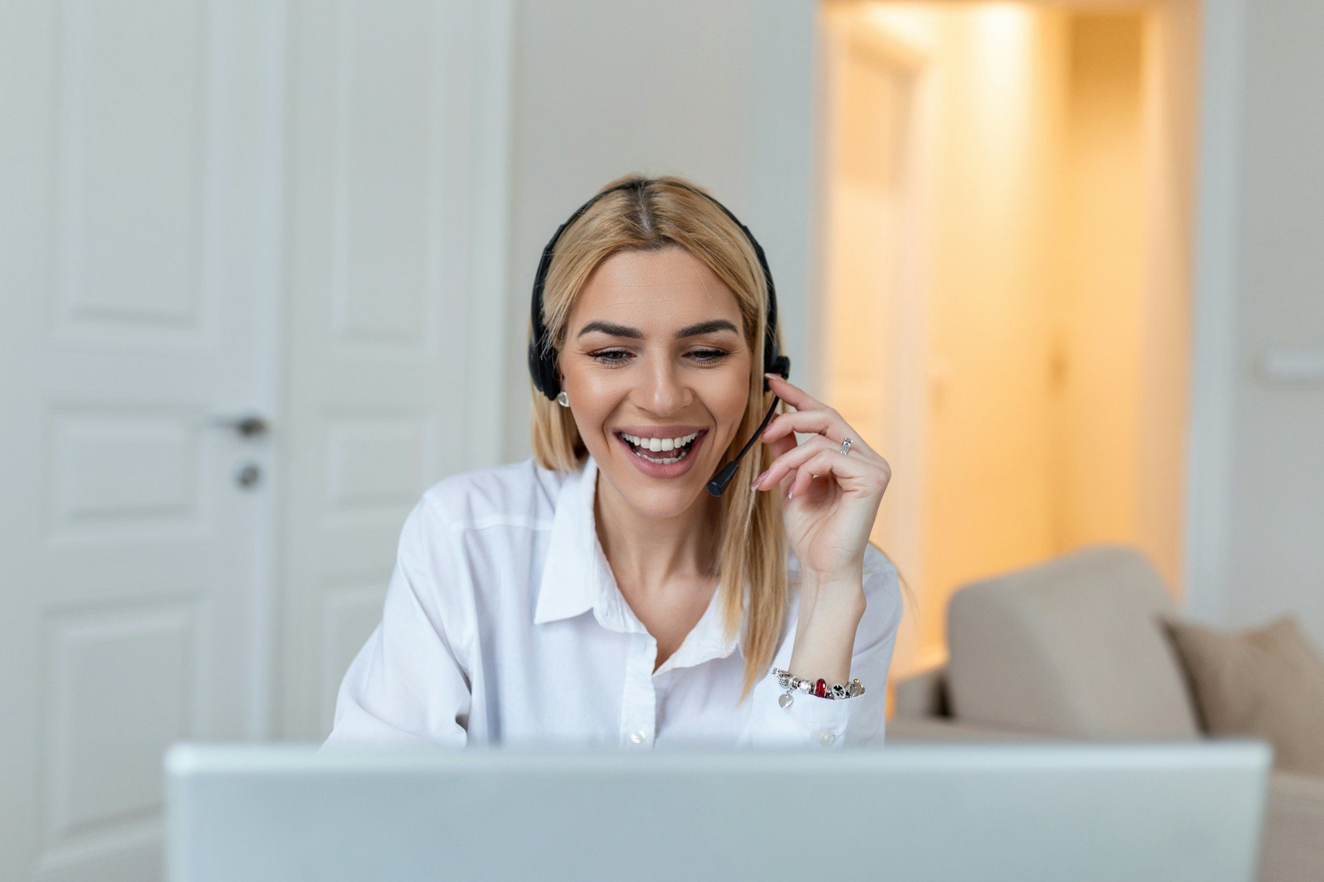 A woman with blonde hair wearing a headset, smiling, and talking while looking at a screen.