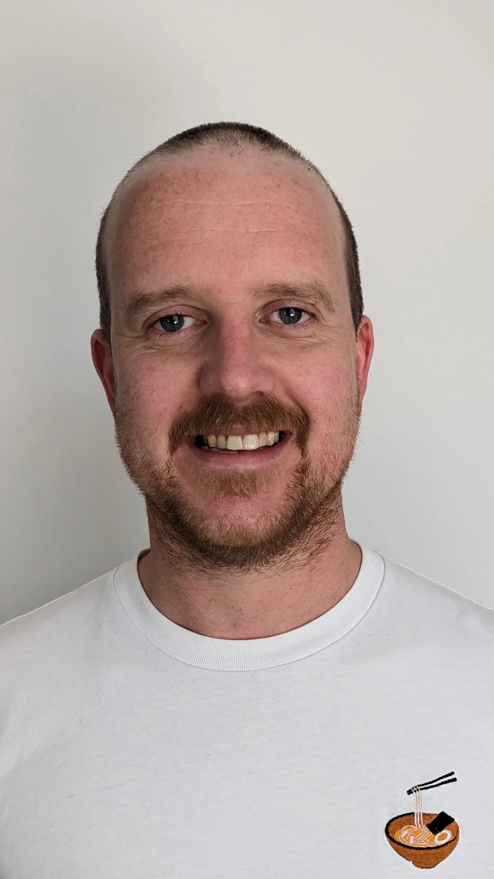 A man with a beard wearing a white t-shirt, smiling at the camera.