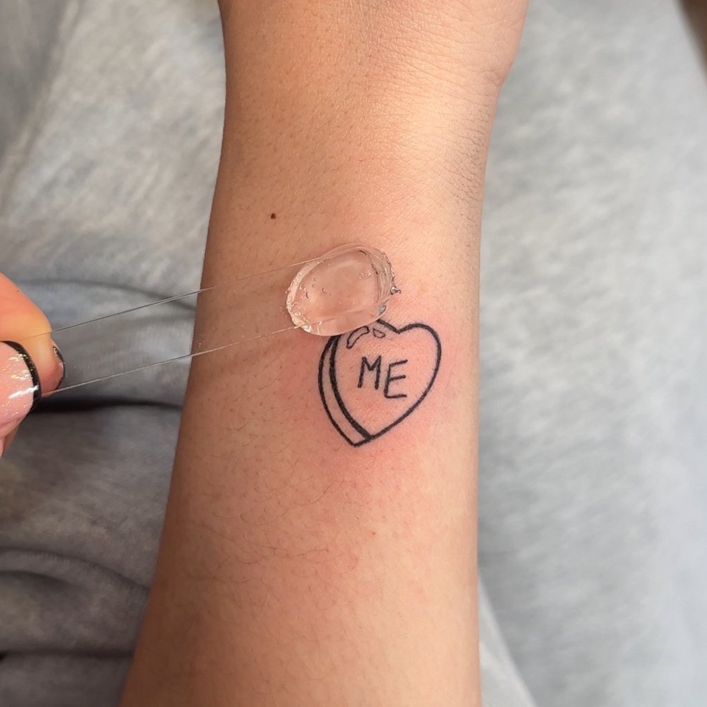 Fine line tattoo of heart with 'ME' and application of ointment on arm in Los Angeles