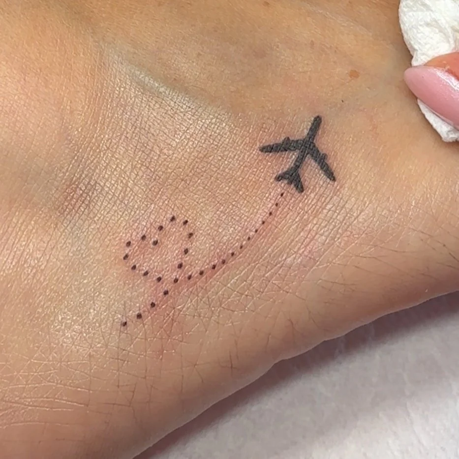 Close-up of a small tattoo featuring a black airplane and a dotted line forming a heart shape on the skin done in Los Angeles
