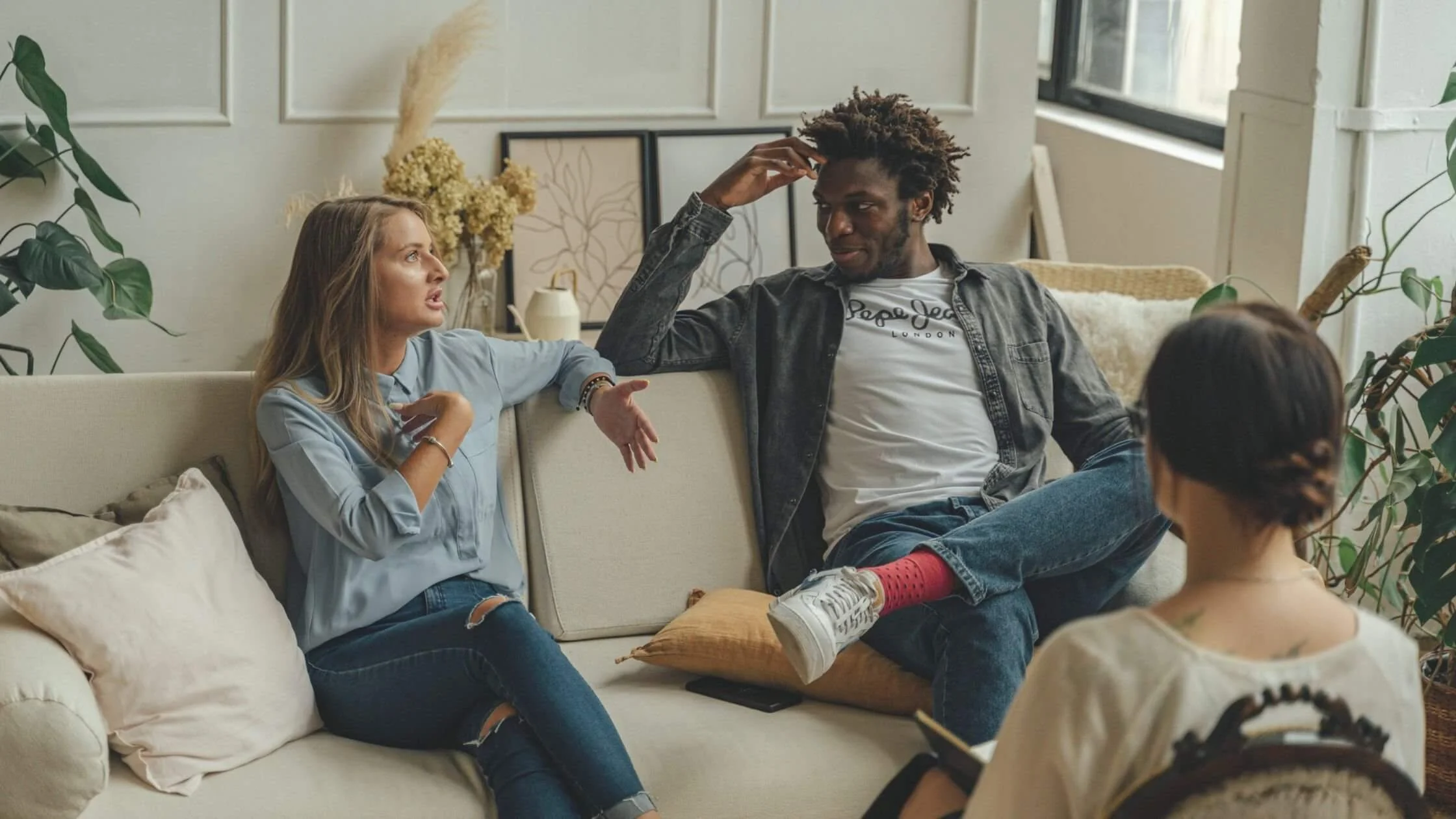 Three people having a serious conversation in a cozy living room with natural light, plants, and artwork in the background.