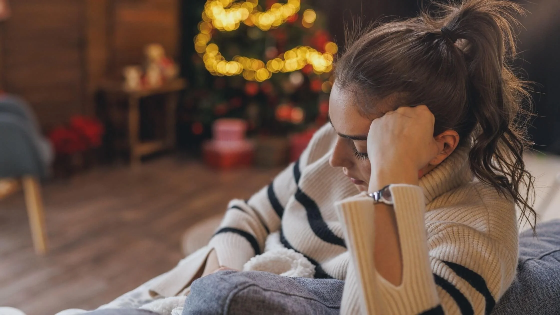 A woman sitting on a couch with her eyes closed and head resting on her hand, in a cozy living room decorated for Christmas with a tree and blurred presents in the background.