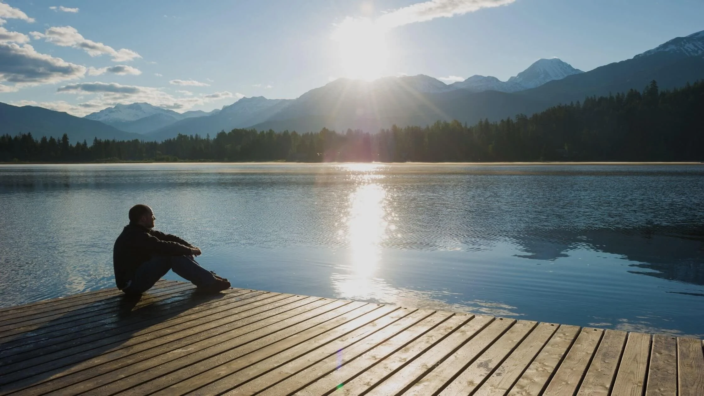 A person sitting on a wooden dock by a lake, with mountains and trees in the background, under a bright sun in the sky.