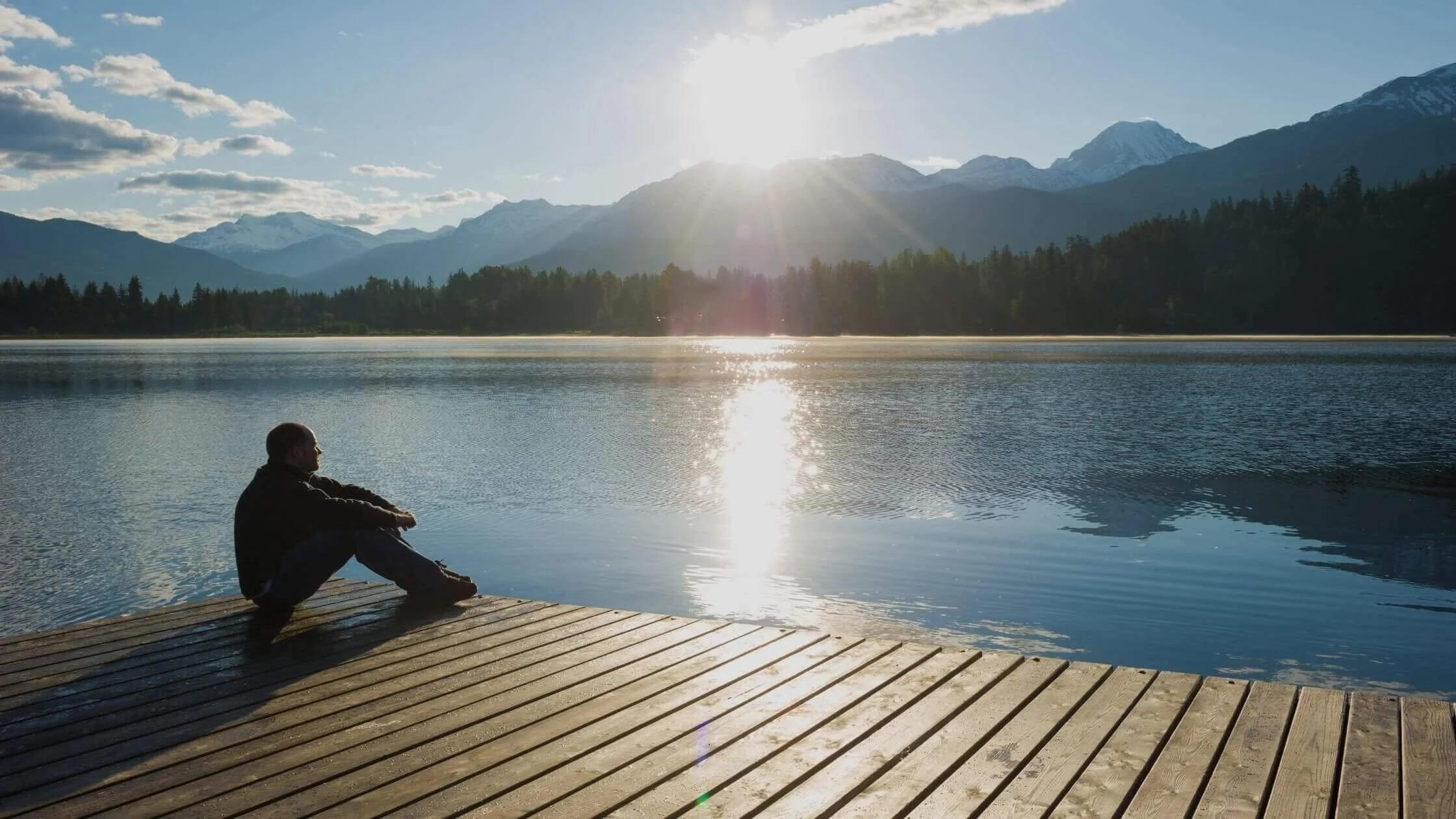 A person sitting on a wooden dock by a lake, with mountains and trees in the background, under a bright sun in the sky.