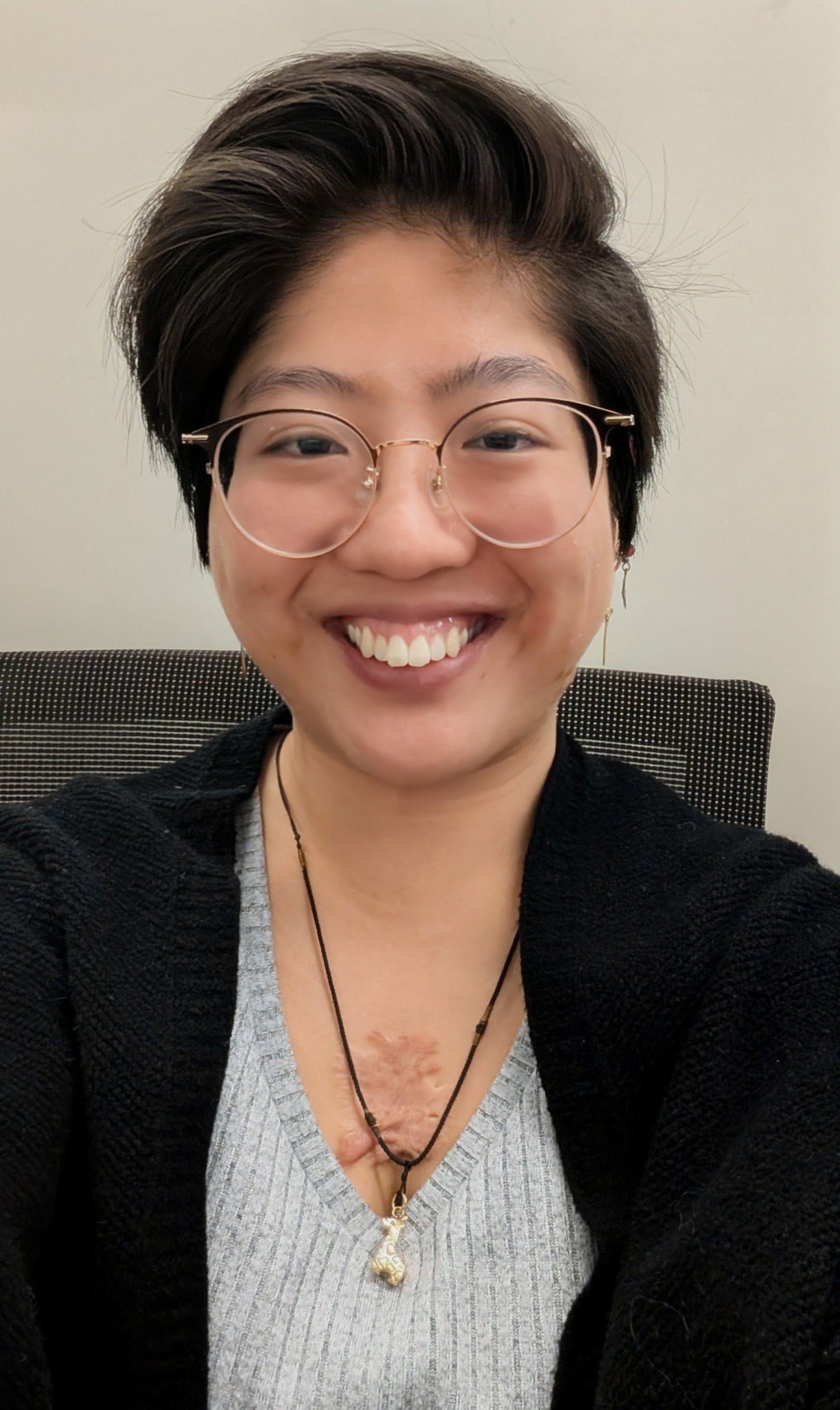 A woman with short dark hair, wearing glasses, a gray top, a black cardigan, and layered necklaces, smiling at the camera while sitting in front of a plain wall.