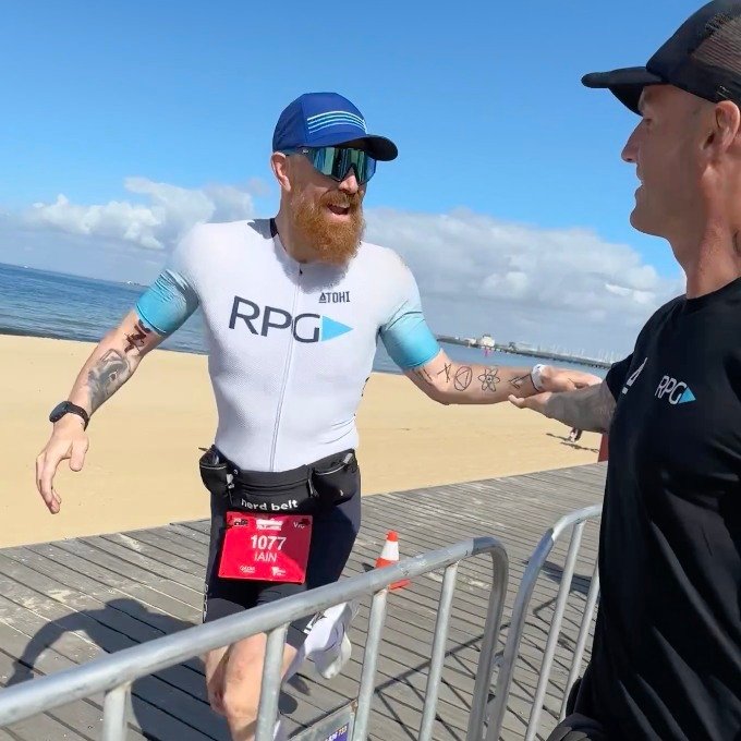 A bearded runner wearing a white “RPG” triathlon suit, sunglasses, and a race bib reaches out to high-five a coach beside a metal barrier on a sunny beachside boardwalk.