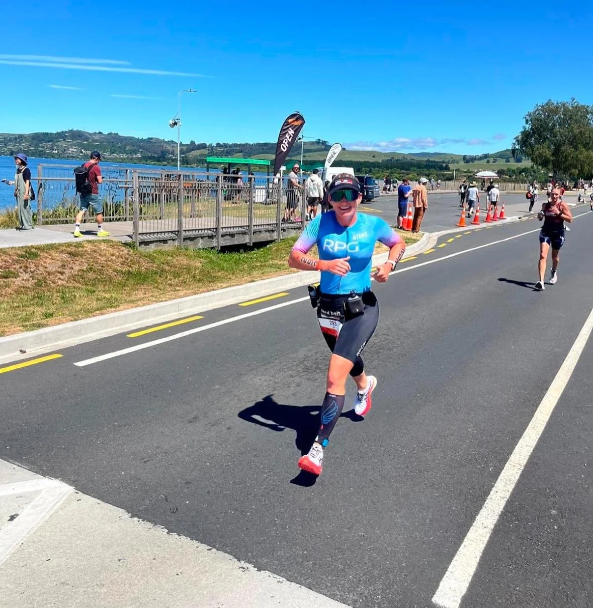 Female athlete in RPG Coaching tri suit running on a sunny waterfront road during a long-course triathlon event.