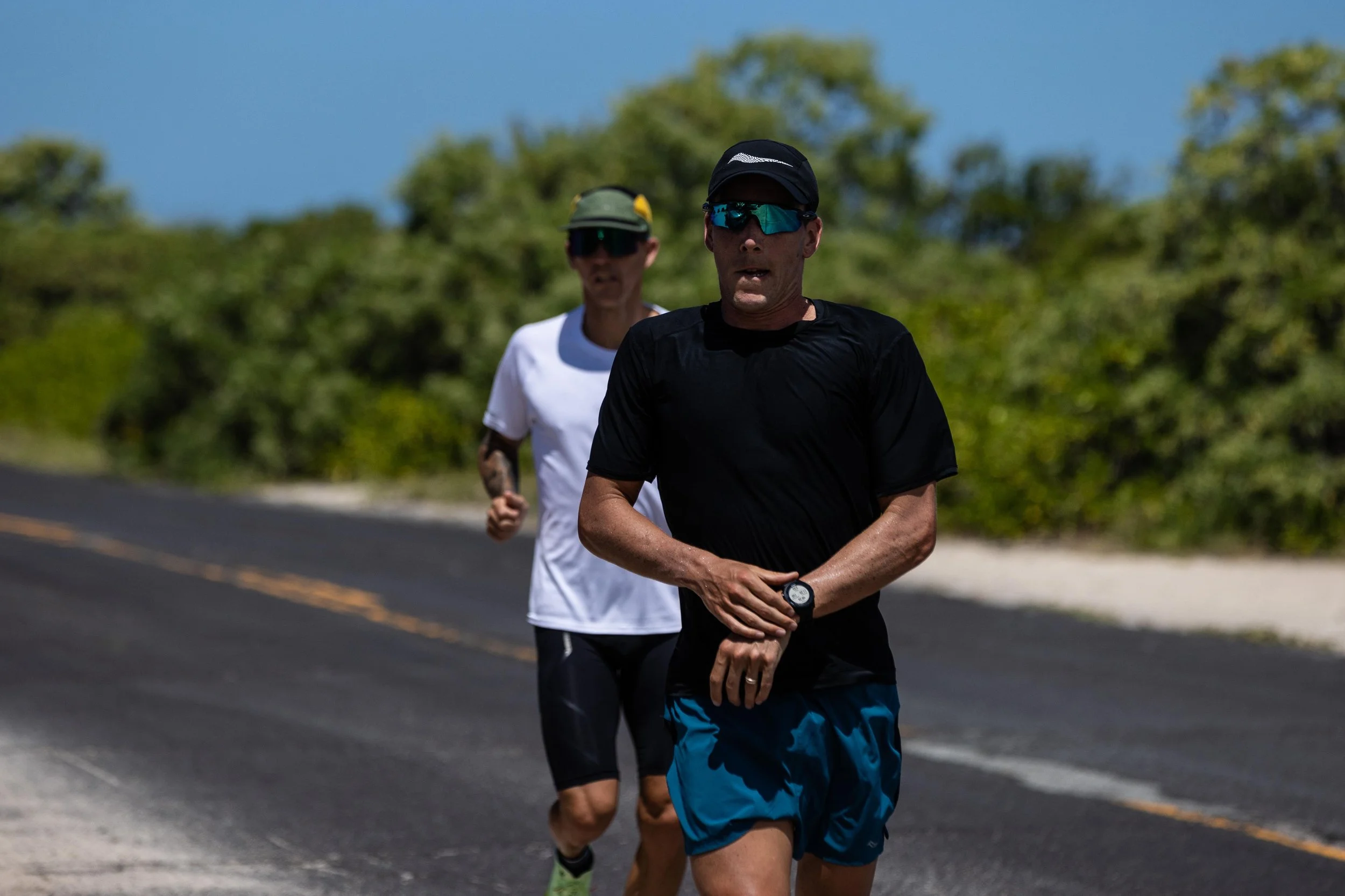 Two runners training on a sunlit road in hot conditions, with the lead runner checking a sports watch during the session.