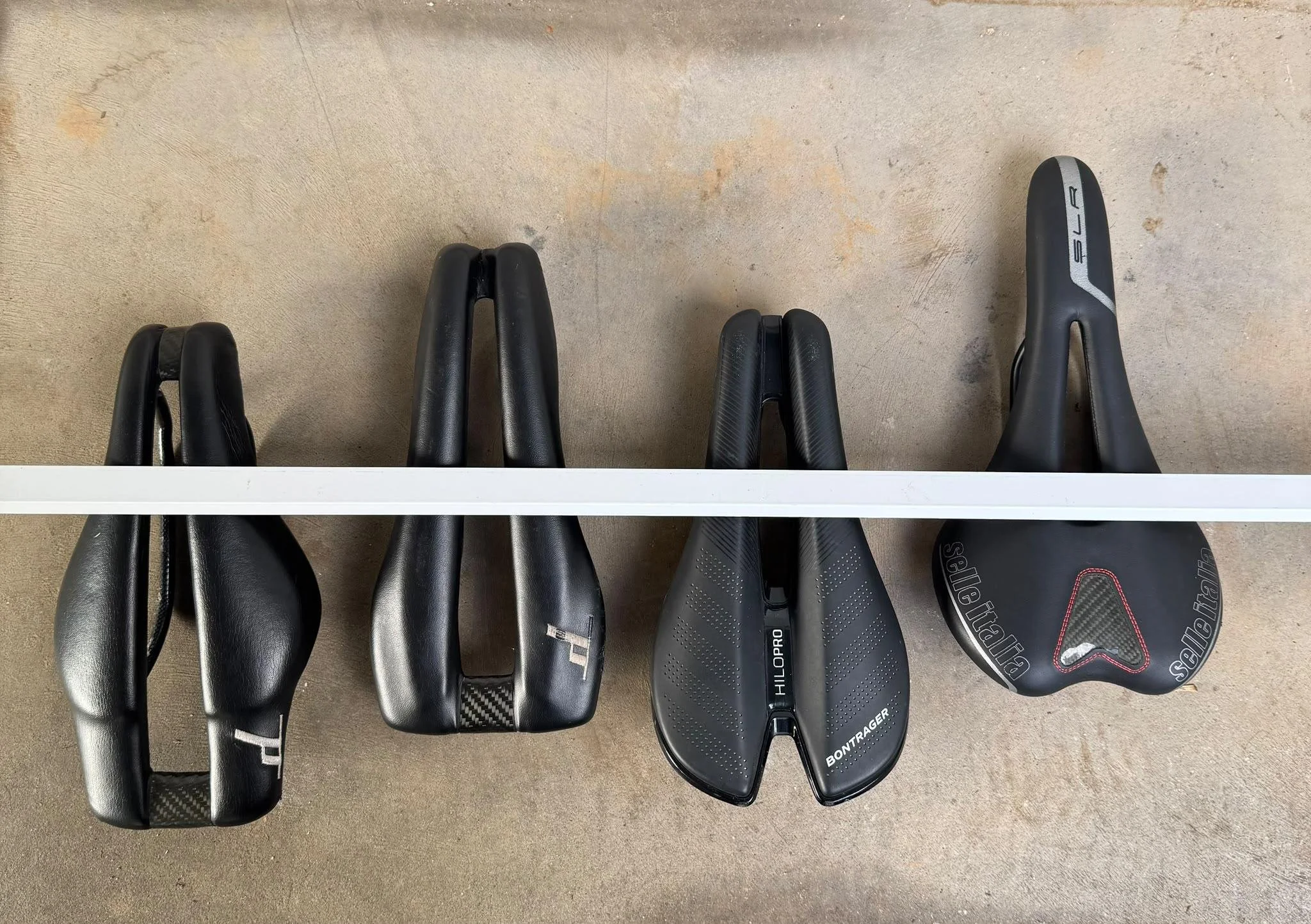 Overhead view of four black split-nose bicycle saddles arranged side by side beneath a white measuring bar on a concrete floor.