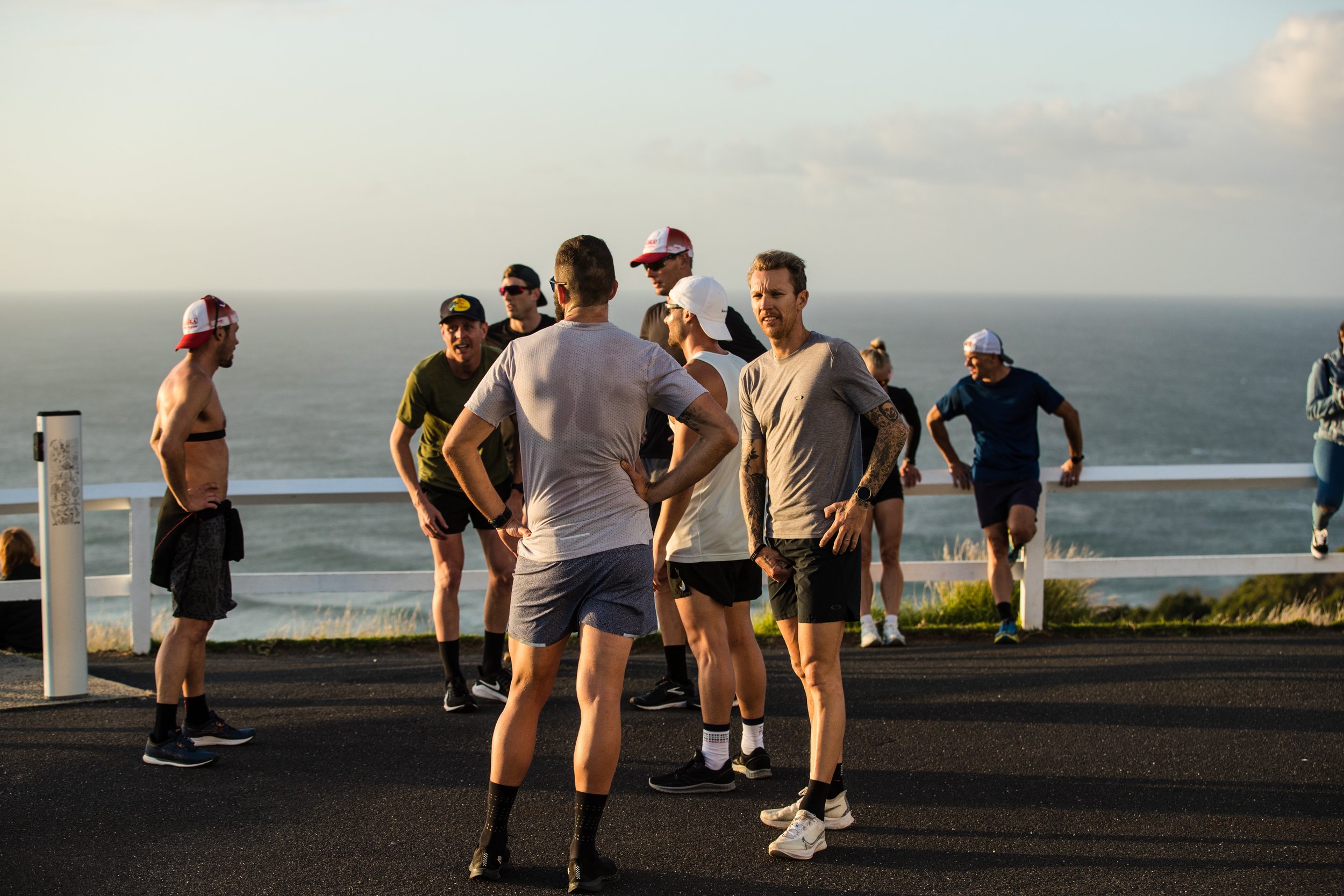 A group of runners in athletic wear stand together on a coastal road, stretching and chatting, with the ocean visible in the background during warm, golden-hour light.