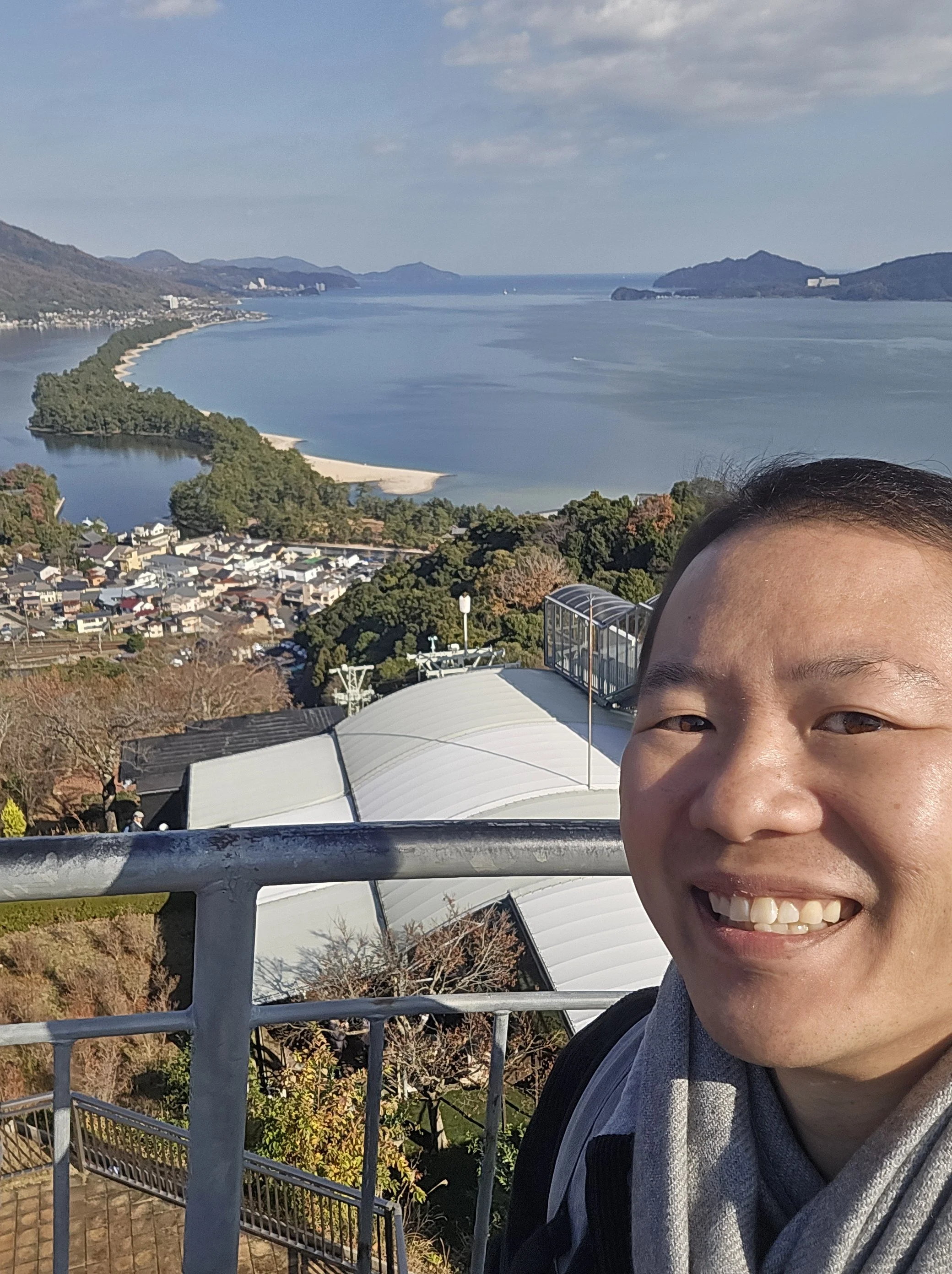 A person smiling with visible teeth, taking a selfie from a high vantage point overlooking a scenic bay with mountains, trees, and a small town below. The person's face is partially visible on the right side of the image.