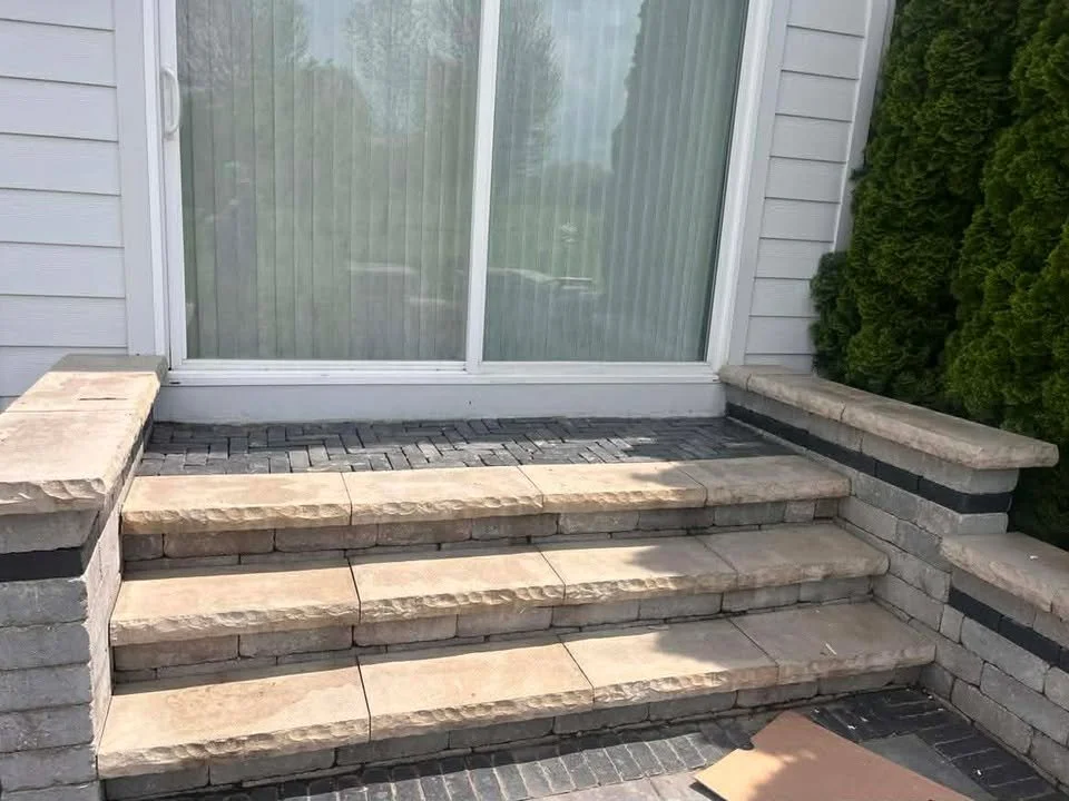 Stone steps leading up to a closed sliding glass door on a house with gray siding and green bushes on the side.