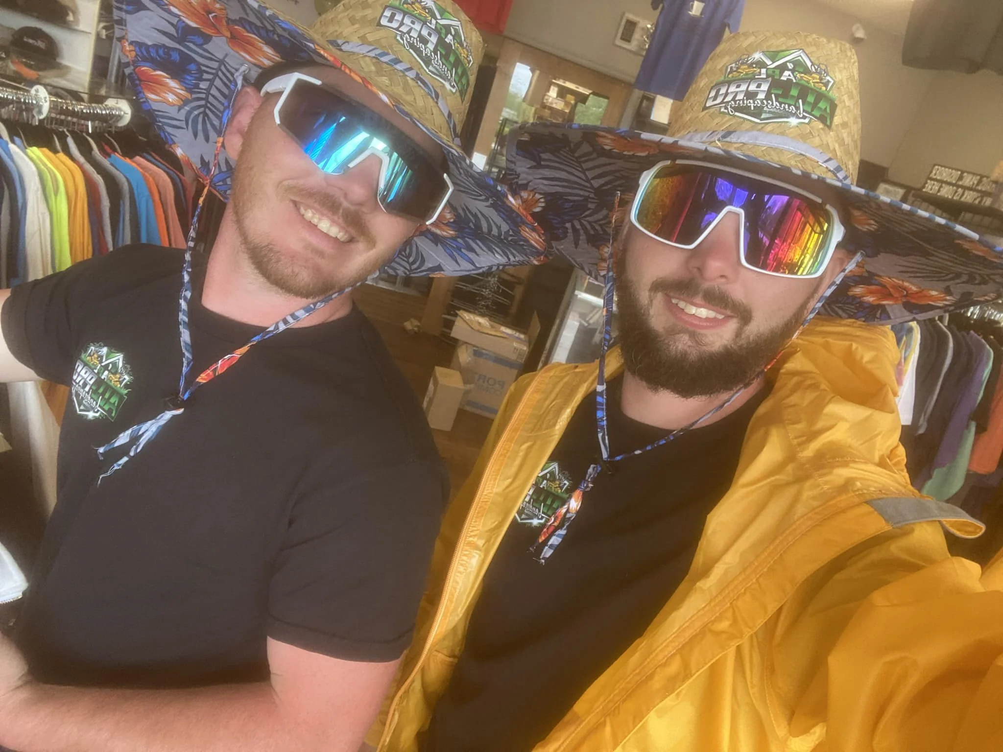 Two men wearing colorful straw hats with a logo, sunglasses, and black T-shirts are smiling and posing for a selfie inside a clothing store. One wears a yellow jacket, and the other has his arm extended for the photo.