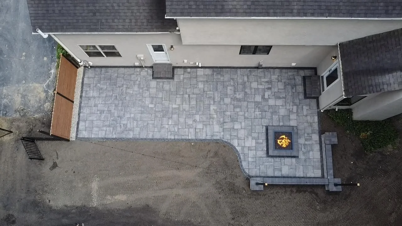 A newly paved backyard patio with gray concrete pavers, a built-in fire feature, and stairs leading up to the house. The yard is bordered by the house on two sides, with an unfinished dirt area in the foreground.