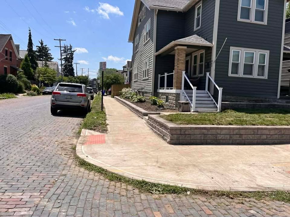Sidewalk with a curb cut accessible ramp leading to a residential building with stairs, grass, and potted plants. Parked cars on a cobblestone street under a partly cloudy sky.