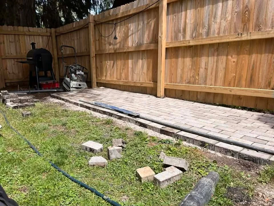 A backyard with a wooden fence, a paved brick walkway, and construction materials including bricks and a drainage pipe, indicating ongoing patio or walkway construction.