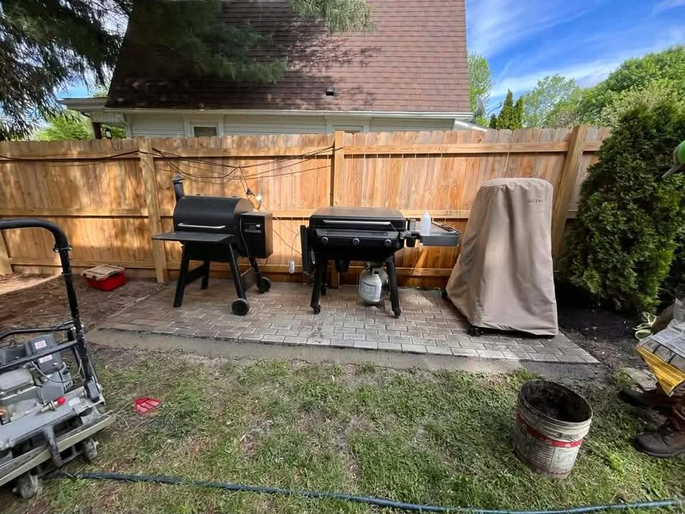 Backyard with a wooden fence, two black grills, covered equipment, a small lawn, and some gardening supplies.