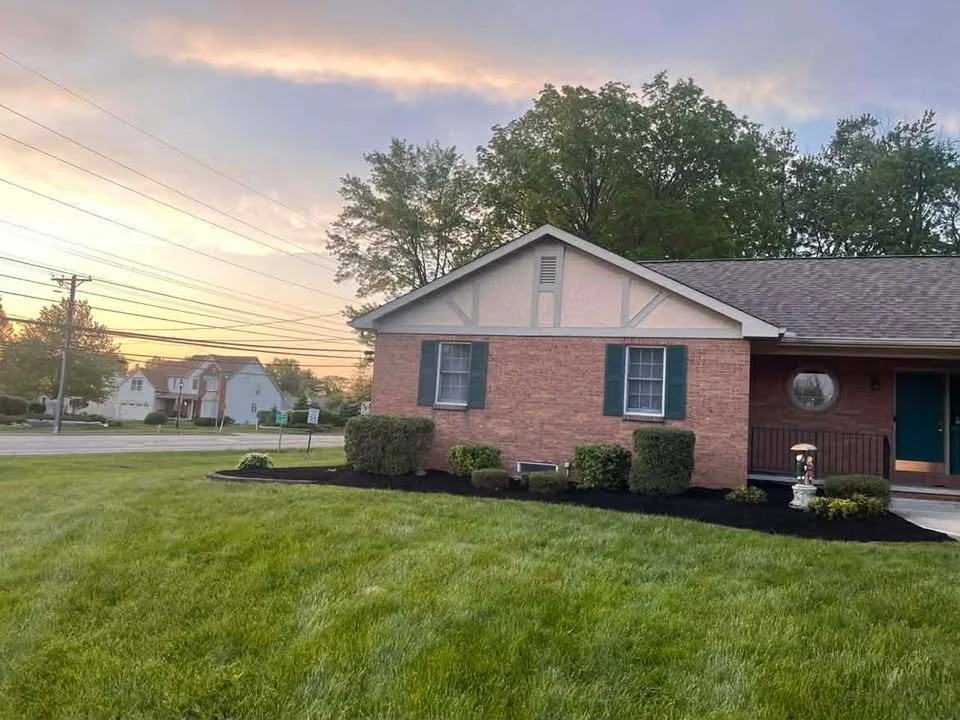 A suburban house with a brick and wood exterior, green shutters, and a landscaped front yard with trimmed bushes and a circular flower bed, at sunset.