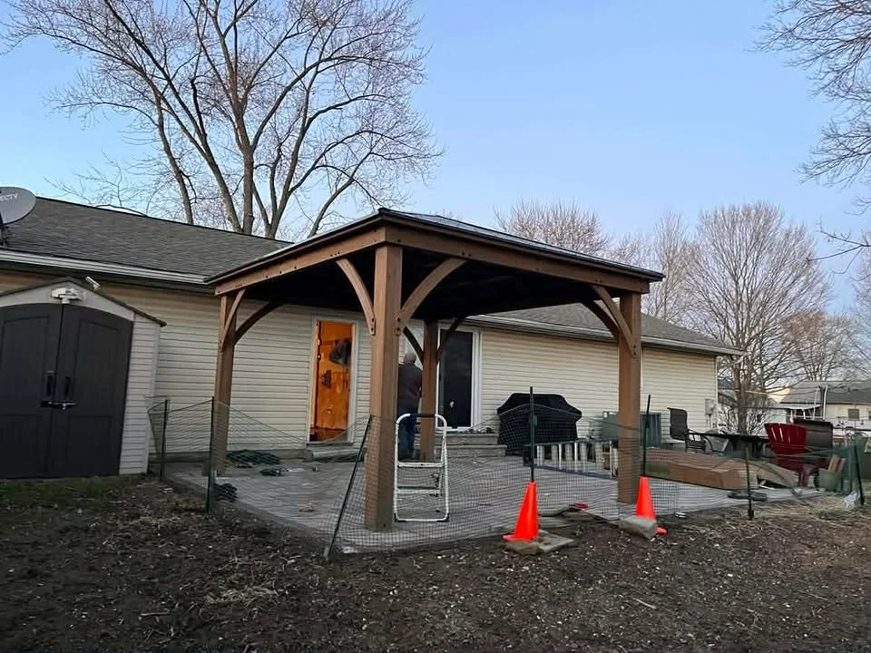 A backyard patio area with a wooden pergola, fencing, and outdoor furniture, on a cloudy day with leafless trees in the background.
