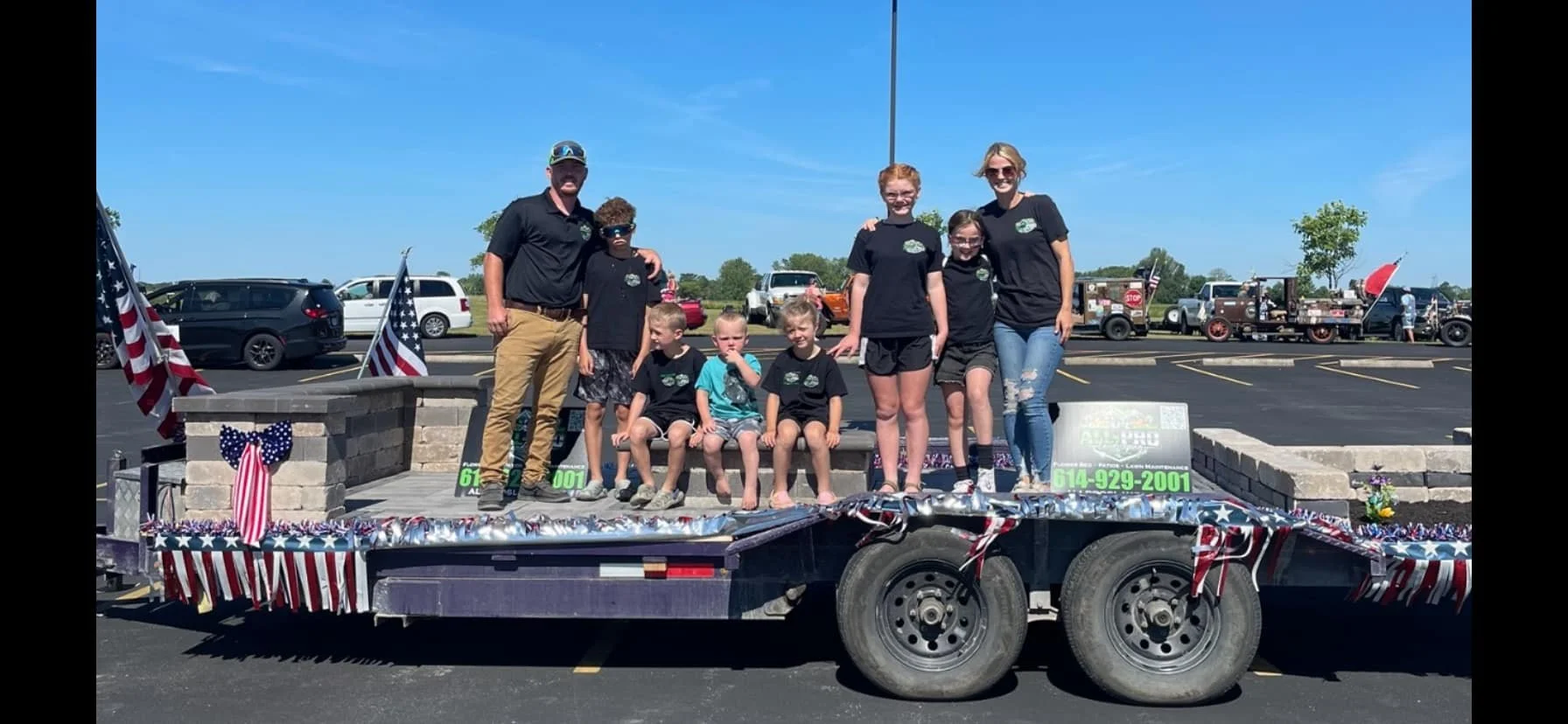 Group of seven children and two adults on a decorated parade float in a parking lot, with American flags, red, white, and blue decorations, and vintage vehicles in the background.