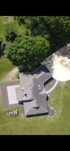 Aerial view of a house with gray roof, surrounded by green trees and lawn, with a paved driveway and outdoor seating area.